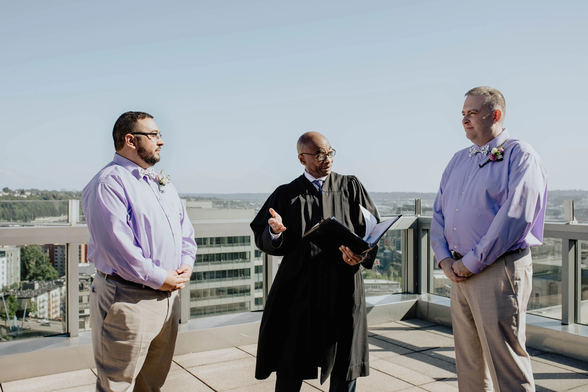 A wedding ceremony on a rooftop with an officiant and two grooms facing each other, city skyline in the background, sunny day. Seattle Municipal Courthouse wedding photography.