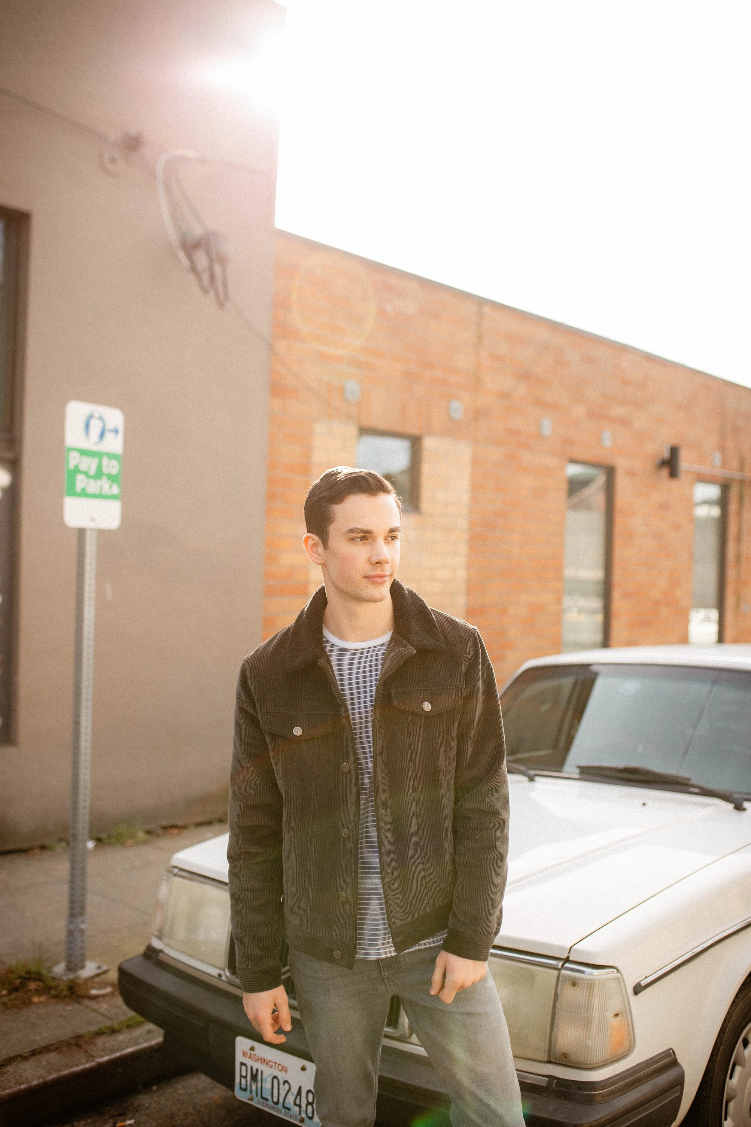 A young man with dark hair in a striped shirt and dark jacket standing near a white car on a city street during daylight. Seattle professional head shot photography