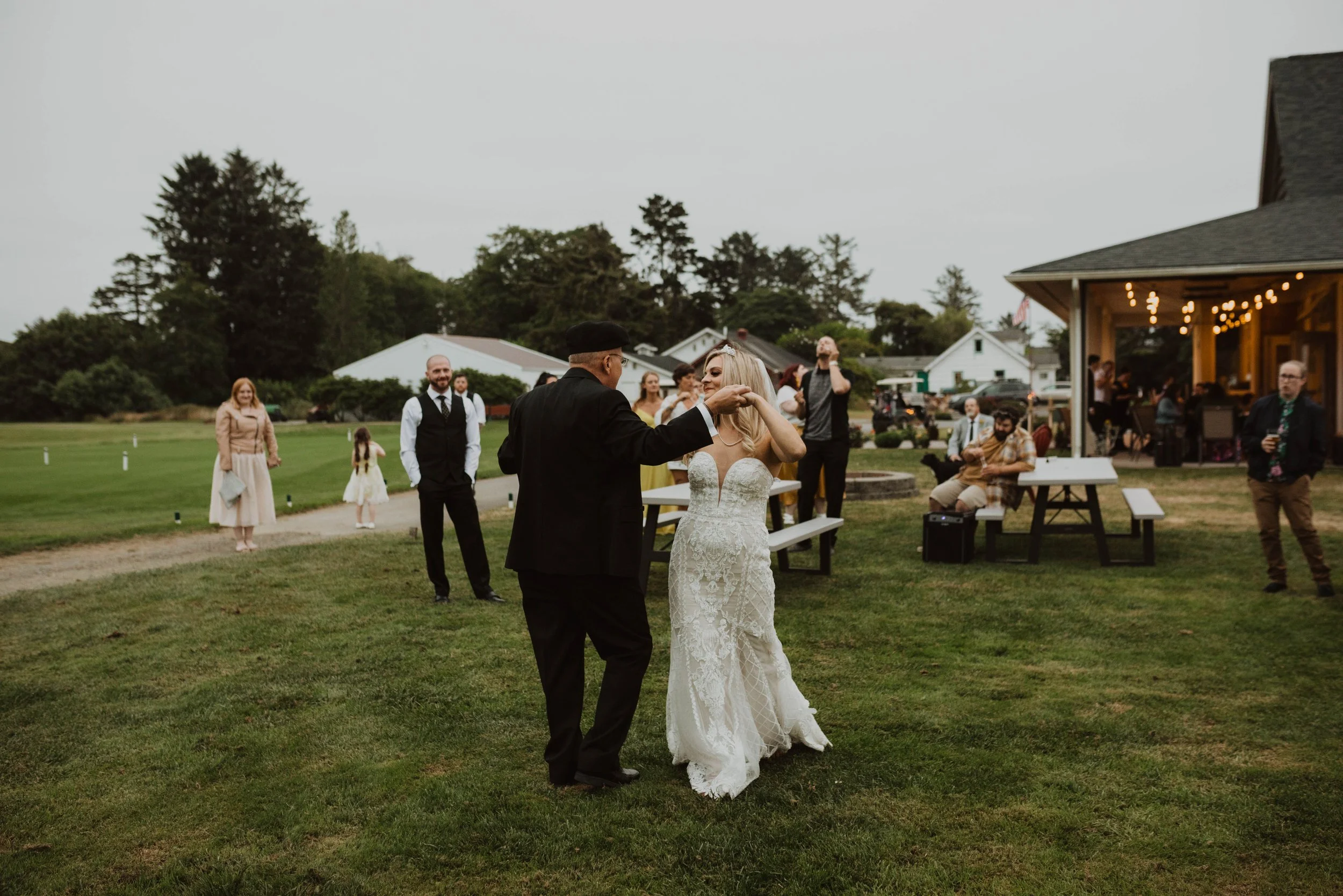 Couple dancing at an outdoor wedding reception during the evening, with guests watching and a rustic building with string lights in the background. Long Beach, WA wedding photography.