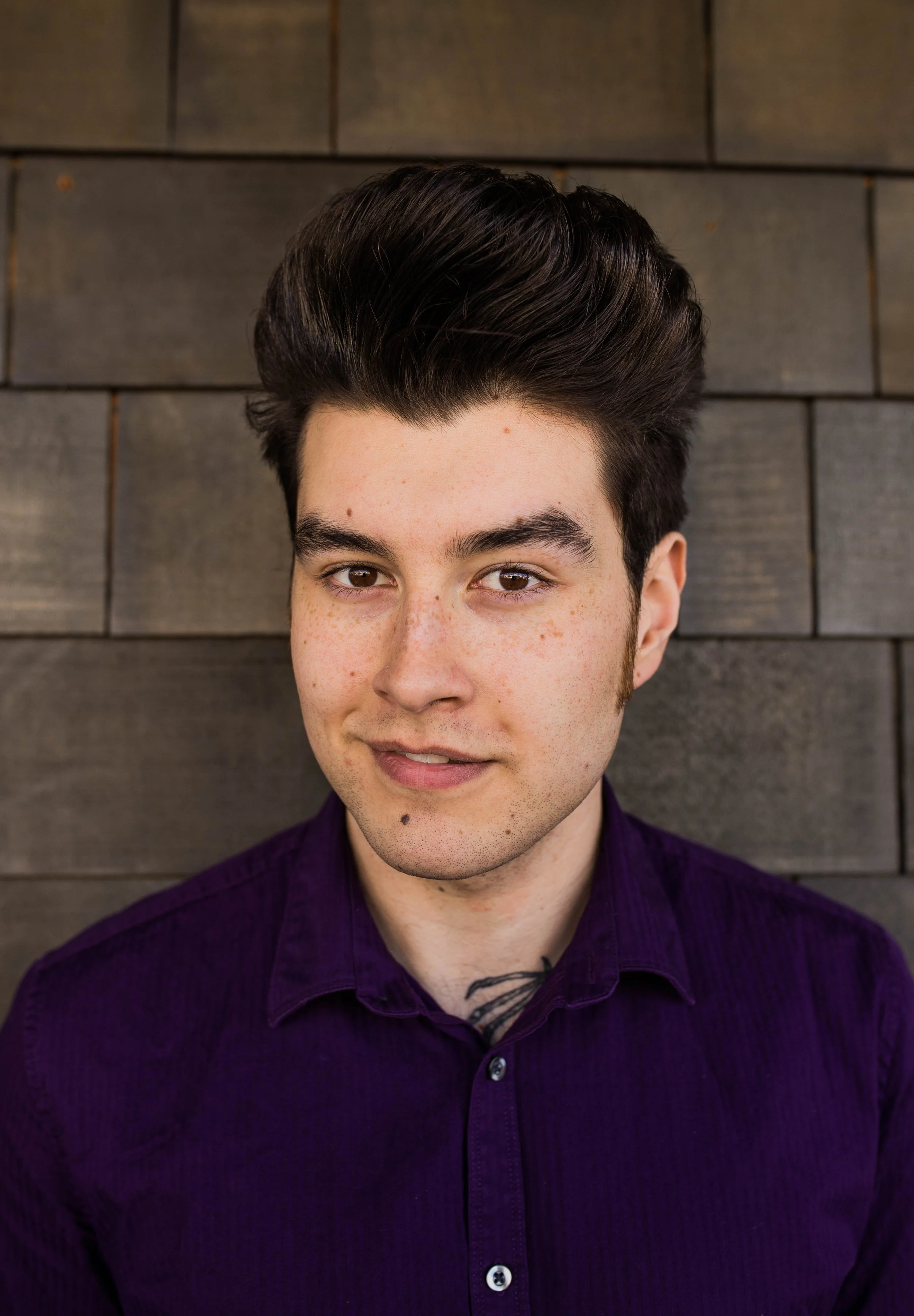 A young man with dark hair and a purple shirt standing against a dark wooden wall. Seattle professional head shot photography