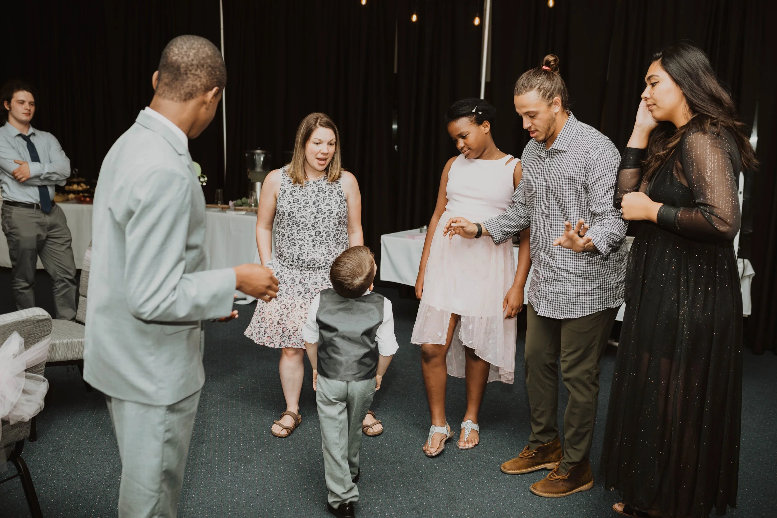 Group of adults and a child dancing together at a social event or party in a room with black curtains and tables in the background. Seattle, WA wedding photography.