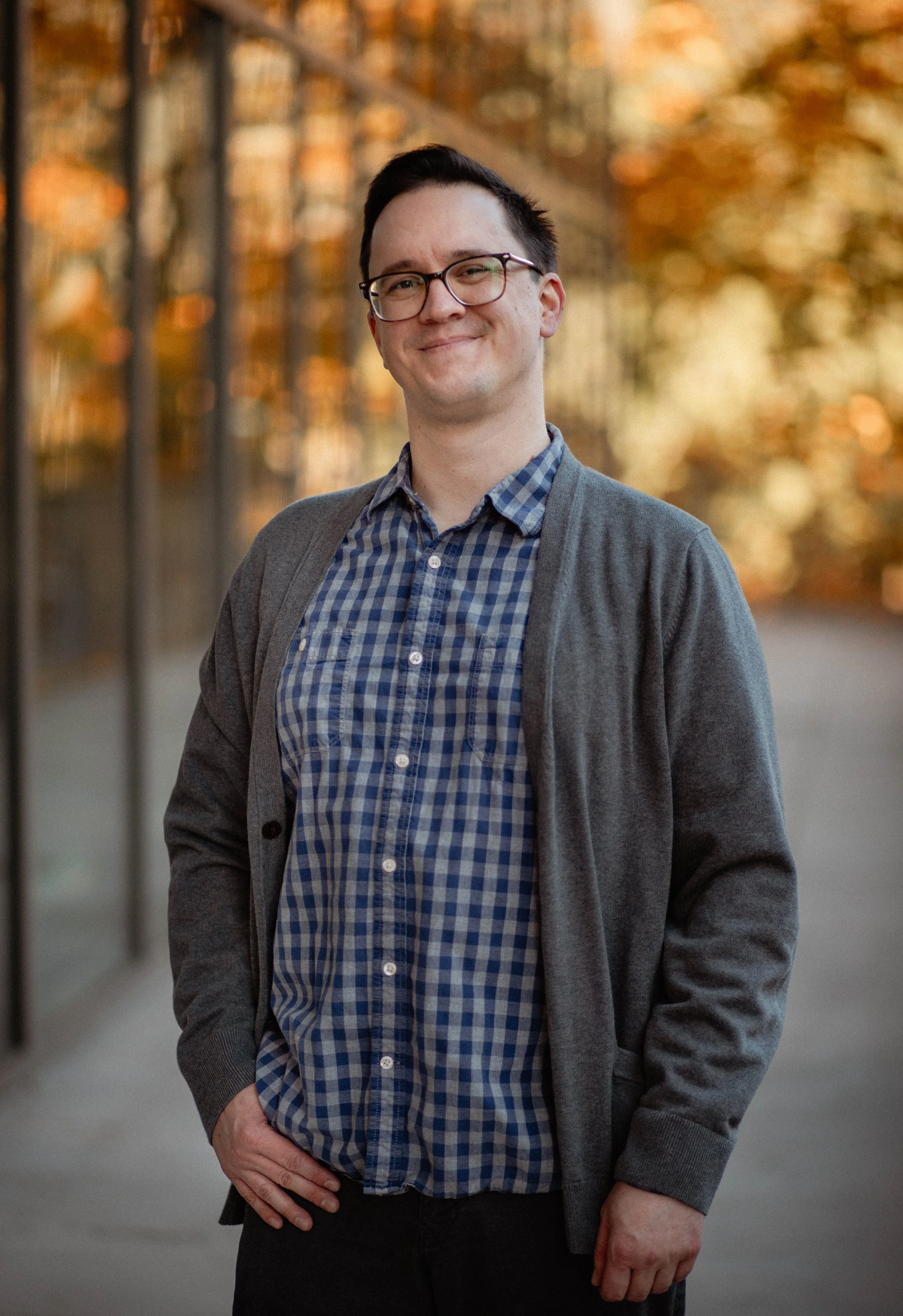 A smiling man with glasses, wearing a gray cardigan and blue checkered shirt, standing outdoors during sunset with autumn-colored trees in the background. Seattle professional head shot photography