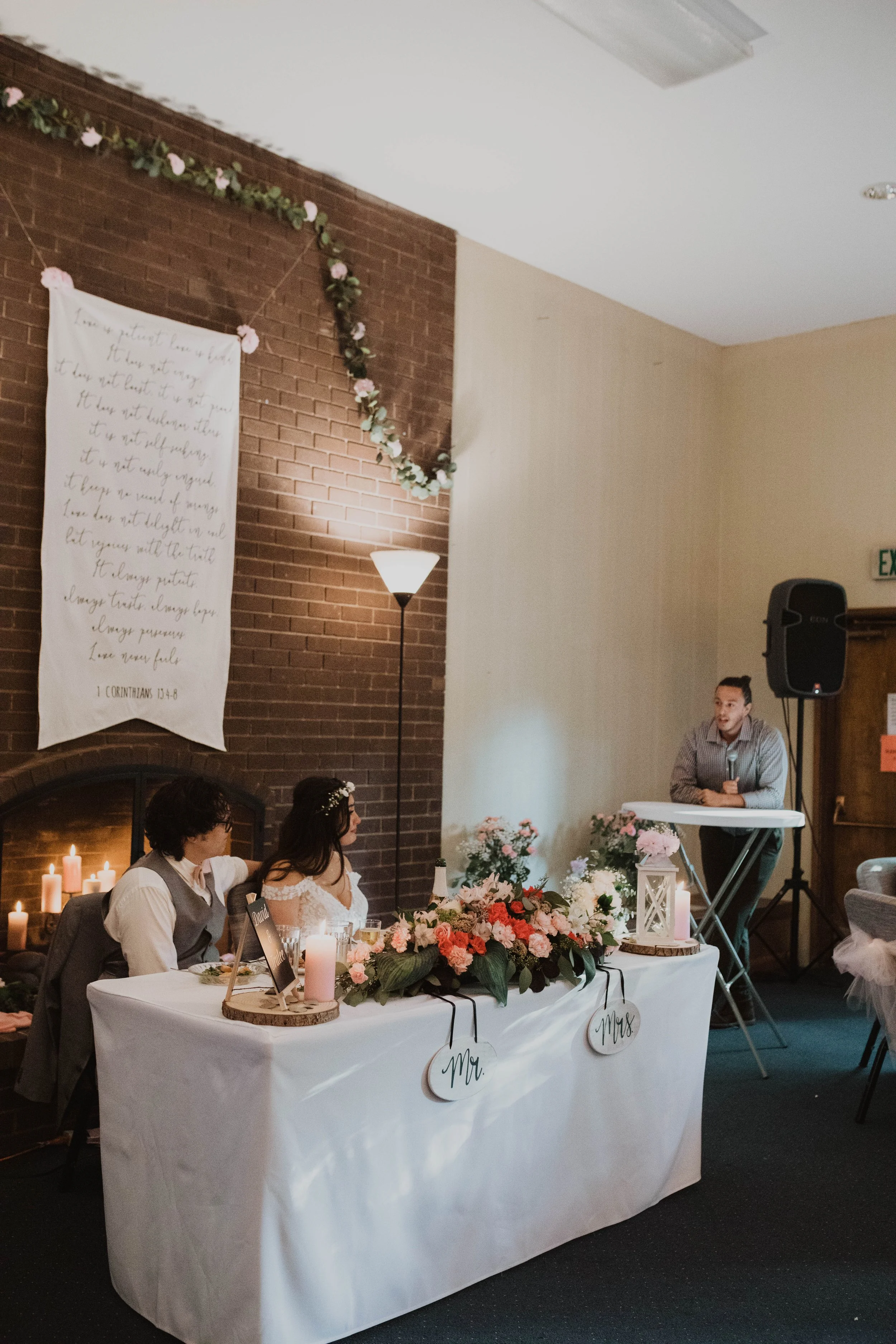 Wedding reception with a bride and groom seated at a table decorated with flowers, candles, and signs reading 'Mr.' and 'Mrs.'; a man stands on the right near a speaker, and a backdrop includes a brick wall, a banner with handwritten text, and floral