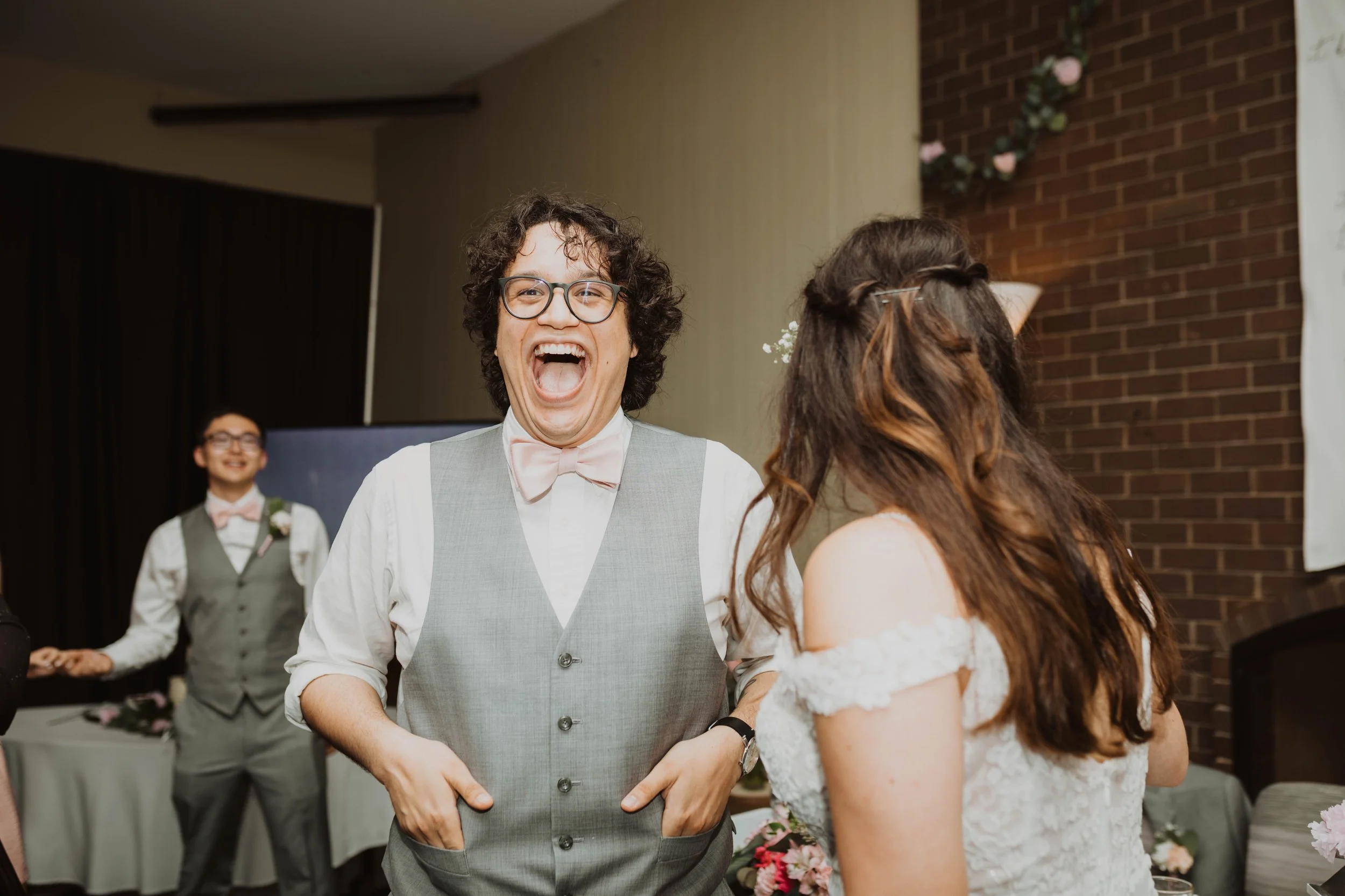 A man with glasses, curly hair, wearing a vest and bow tie, laughing and standing next to a woman with long brown hair and glasses, dressed in a white dress, at a wedding celebration. Seattle, WA wedding photography.