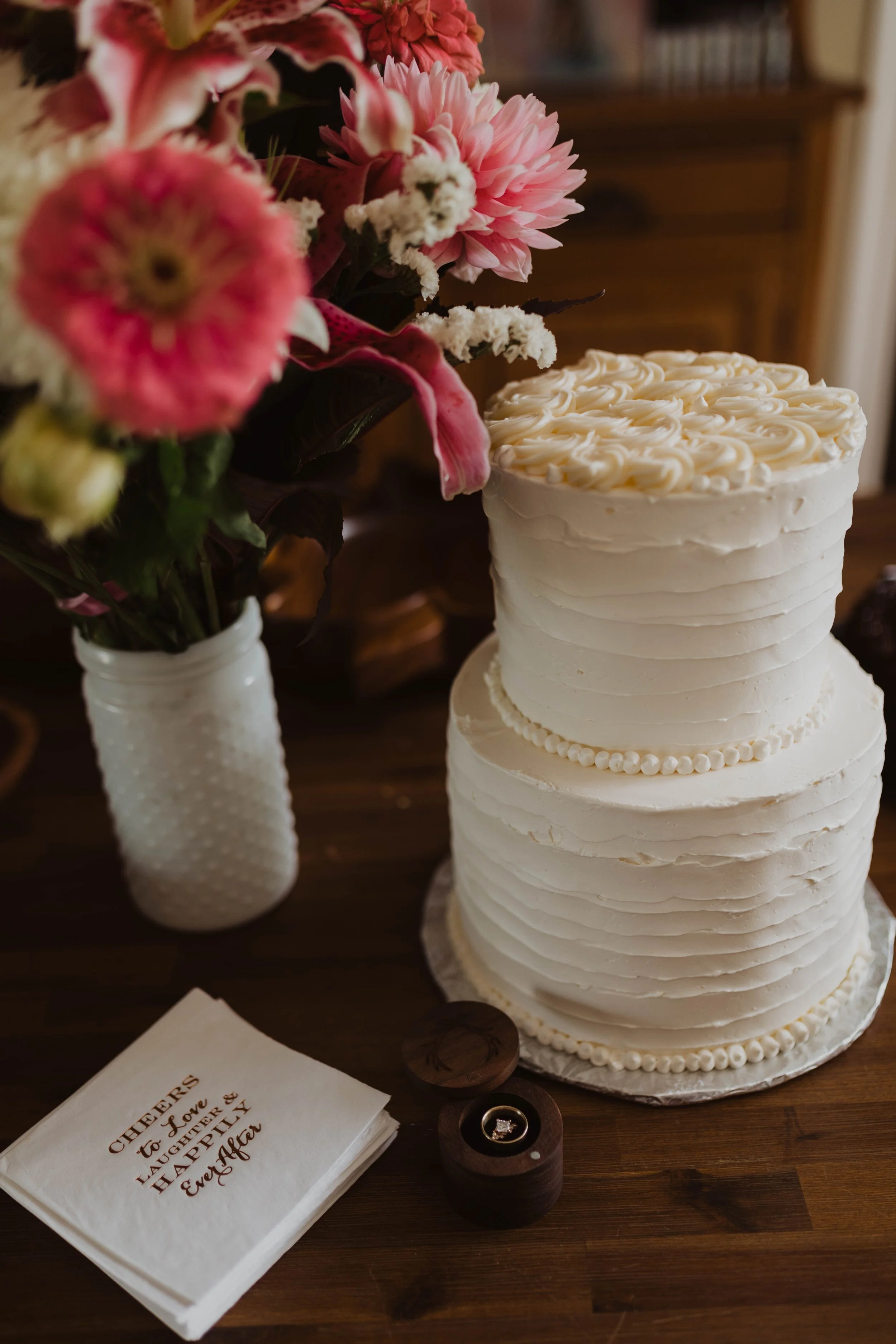 A two-tiered white wedding cake with piped decoration, placed on a wooden table next to a white vase with pink and white flowers, a wedding ring in a wooden box, and a stack of napkins with a heartfelt message. Seattle, WA wedding photography.