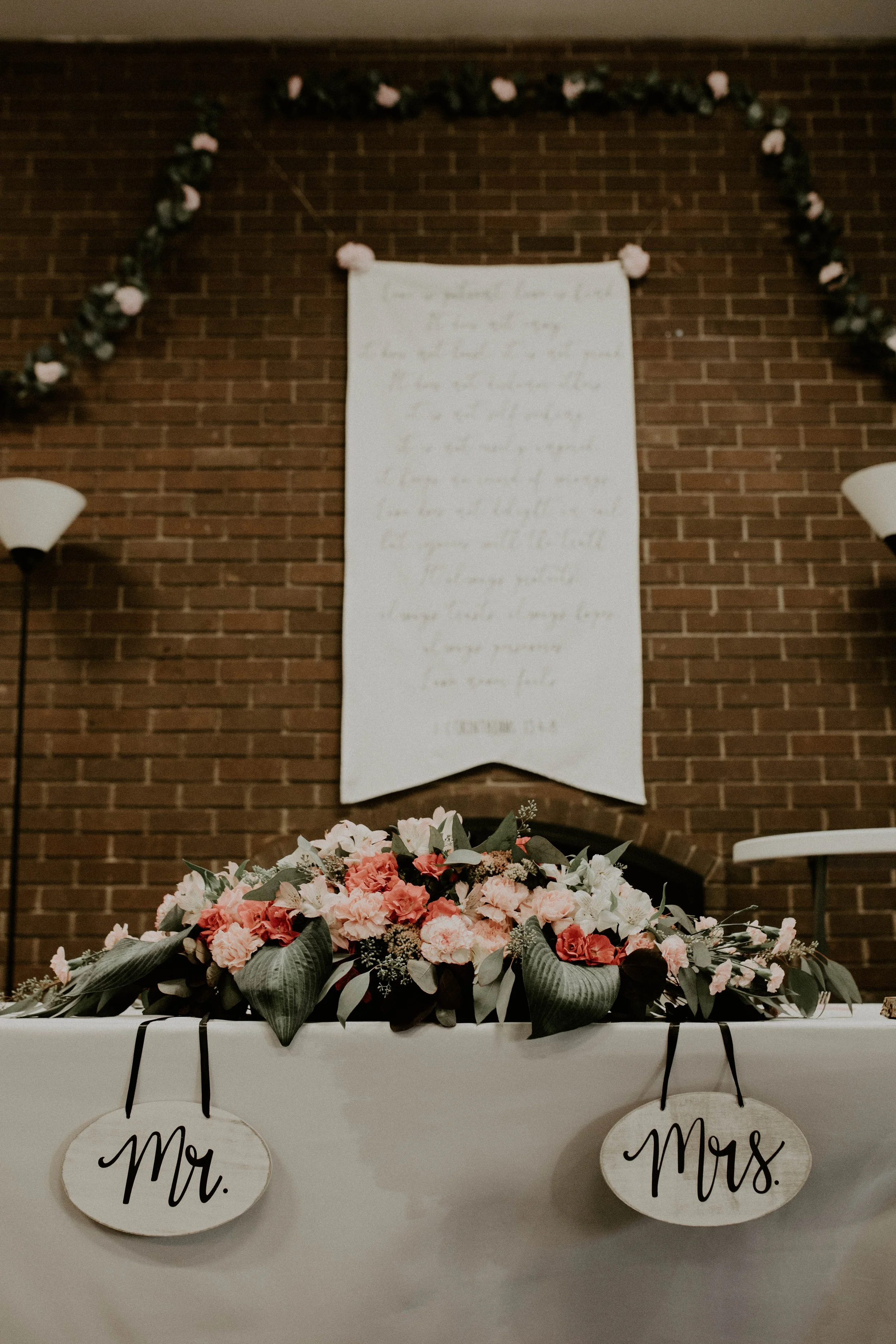 Wedding reception table decorated with pink and white flowers and greenery, with 'Mr.' and 'Mrs.' signs hanging on the front of the table, against a brick wall with a floral garland and a large paper banner in the background. Seattle, WA wedding phot