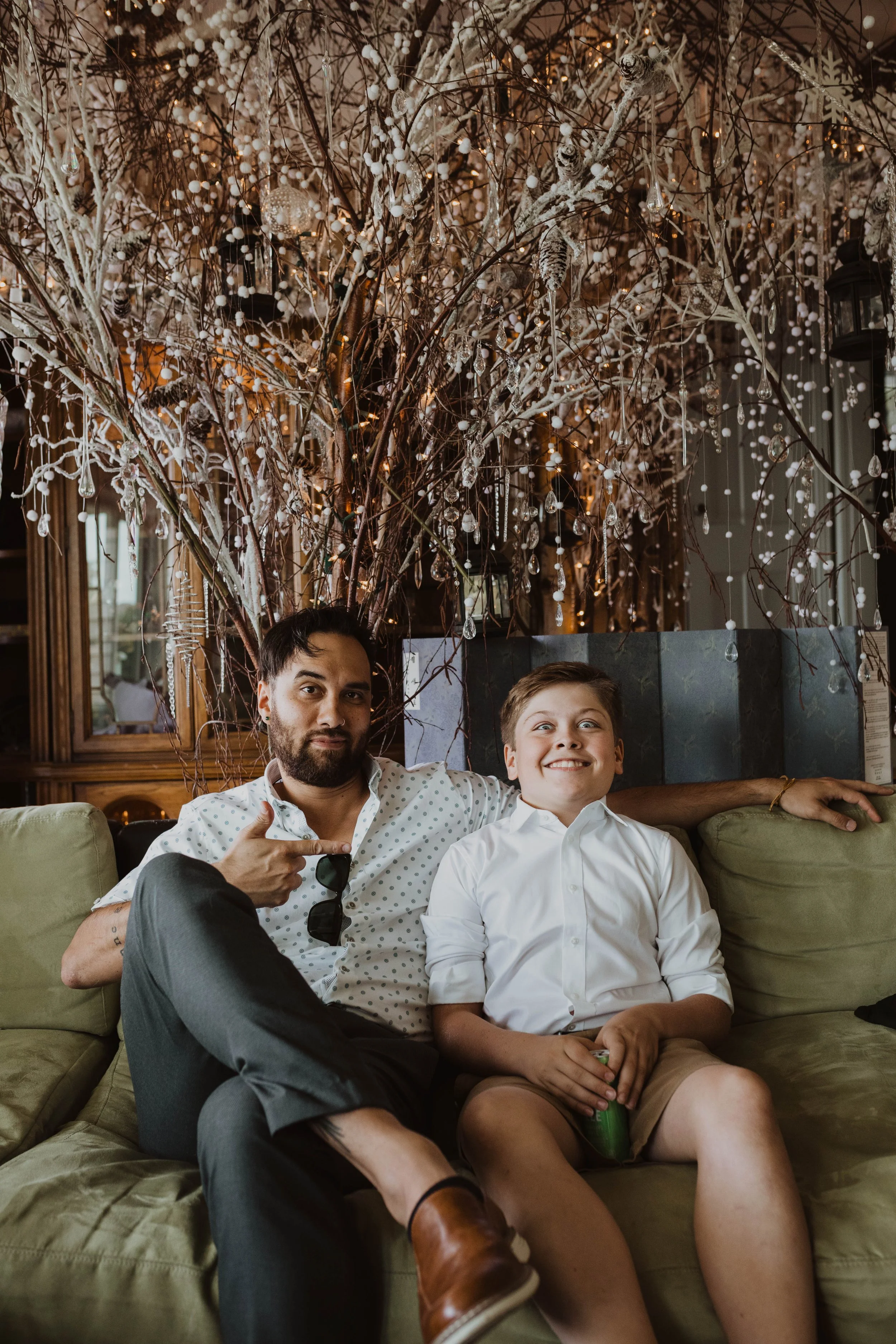 A man with dark hair and a beard sitting on a couch with a young boy. The man is pointing at the boy and the boy is smiling widely. Behind them is an elaborate decorative tree with white branches, beads, and ornaments, and a background of wood paneli
