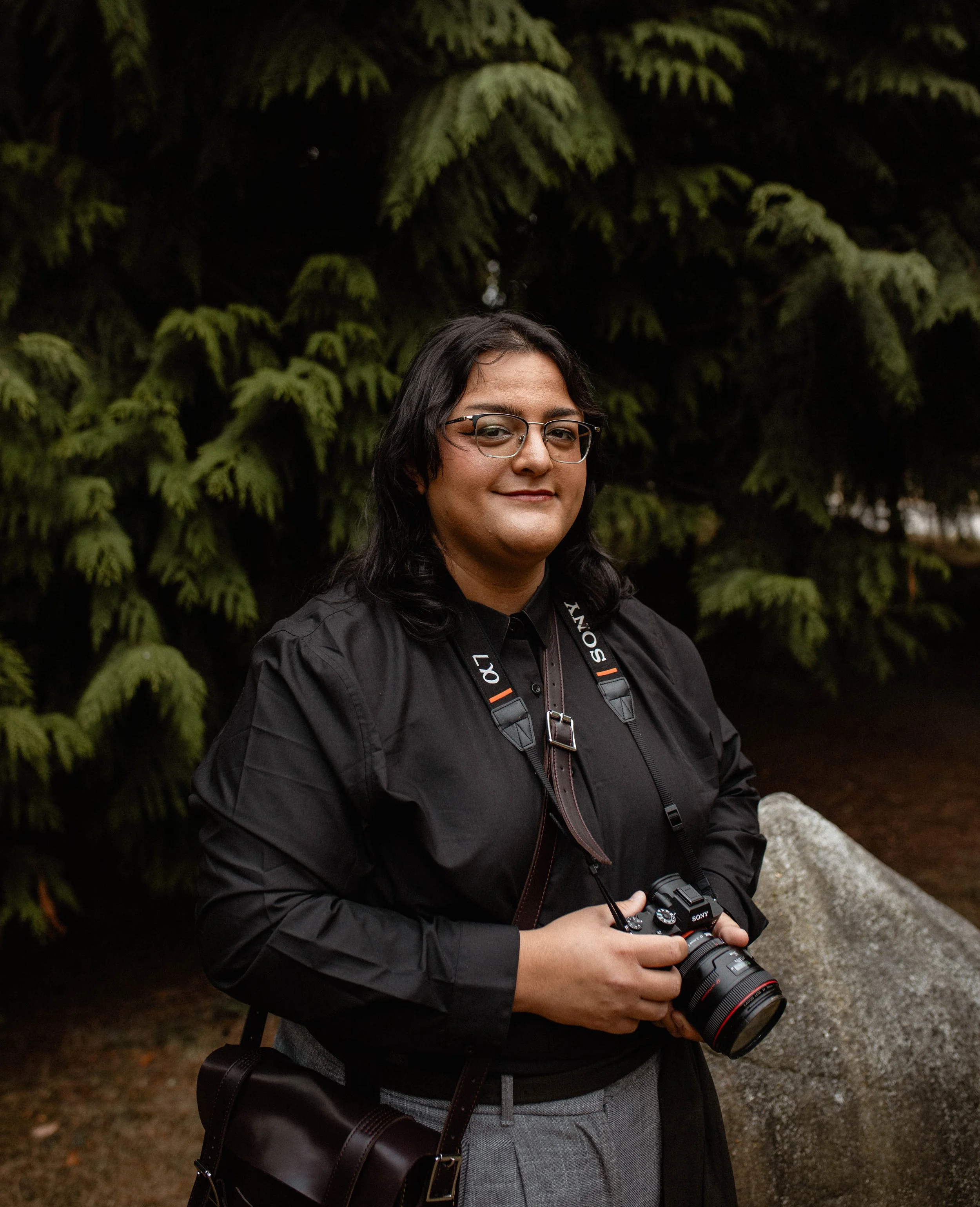 A person with long dark hair and glasses holding a camera, standing outdoors in front of green trees. Seattle professional head shot photography.