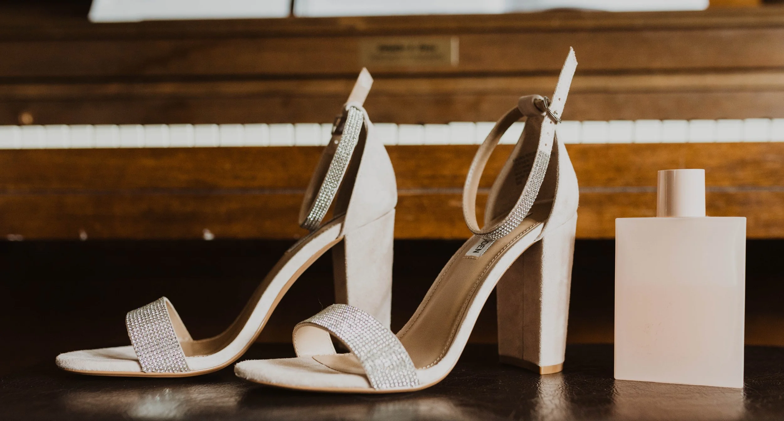 A pair of beige high-heeled shoes with rhinestone embellishments and open toes, placed side by side on a dark surface with a wooden background, and a white perfume bottle beside them. Seattle, WA wedding photography.