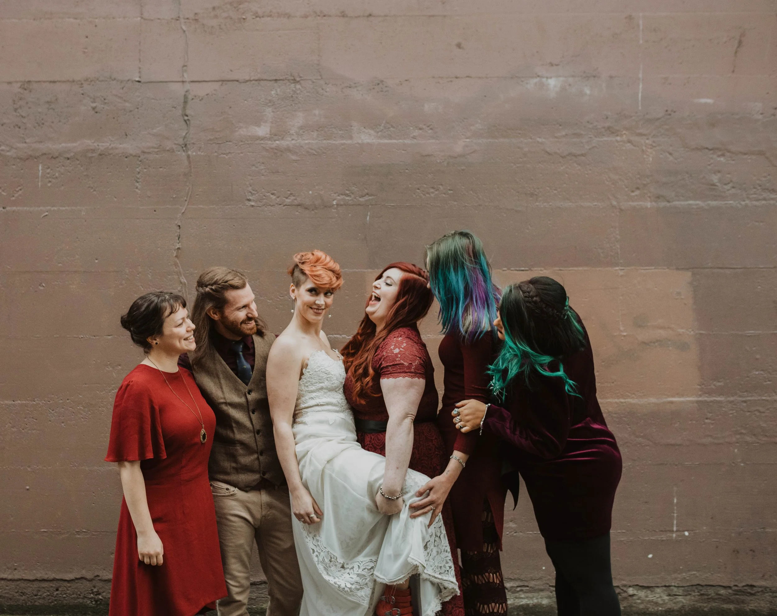 Group of six diverse women and one man, posing together against a plain brick wall. One woman is holding up a bride in a wedding dress, all are smiling and laughing, dressed in colorful, semi-formal attire. Pioneer Square, Seattle, WA wedding photogr