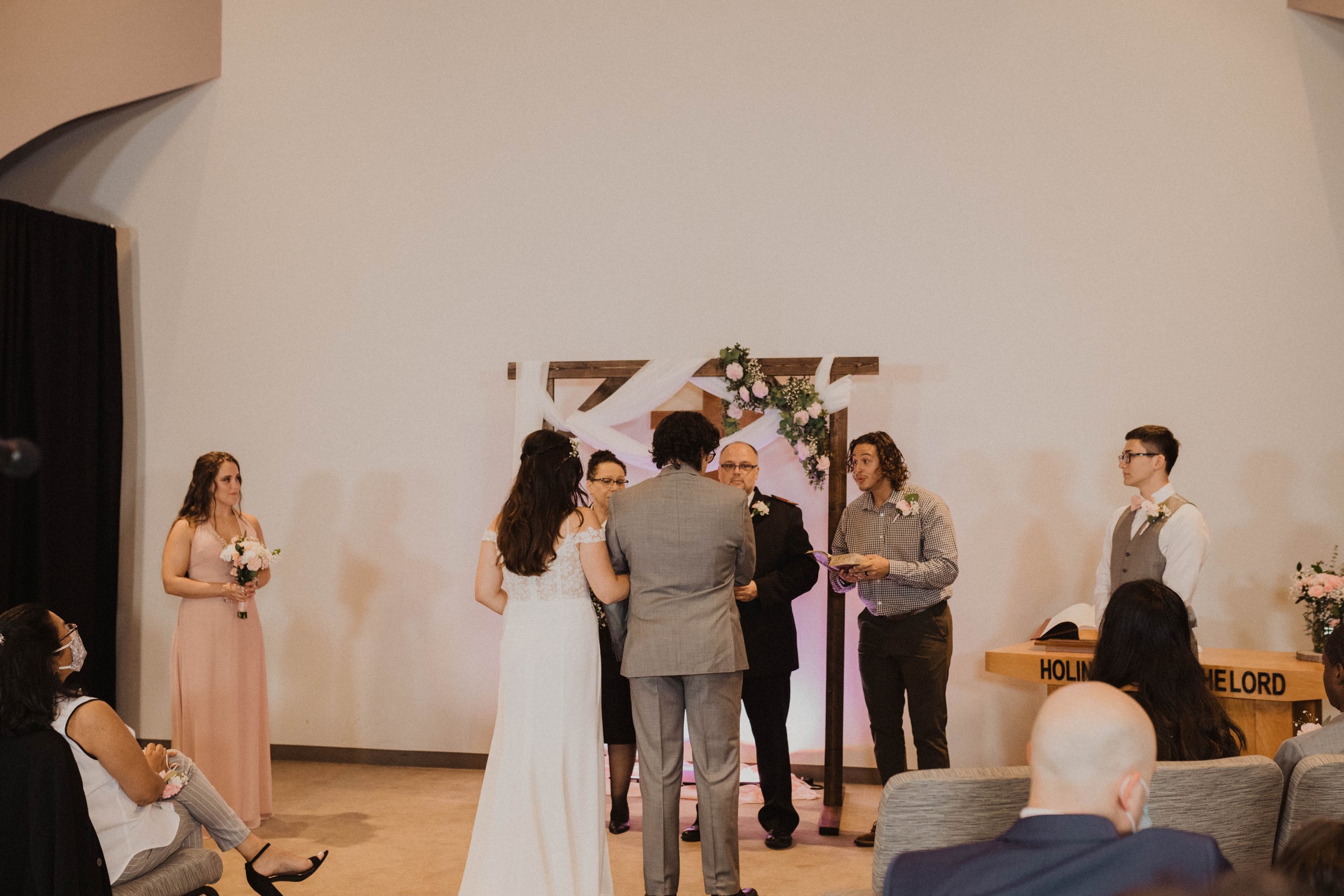 A wedding ceremony with the bride and groom standing before the officiant, surrounded by bridesmaids and groomsmen, inside a decorated venue with a wooden arch and floral arrangements. Seattle, WA wedding photography.