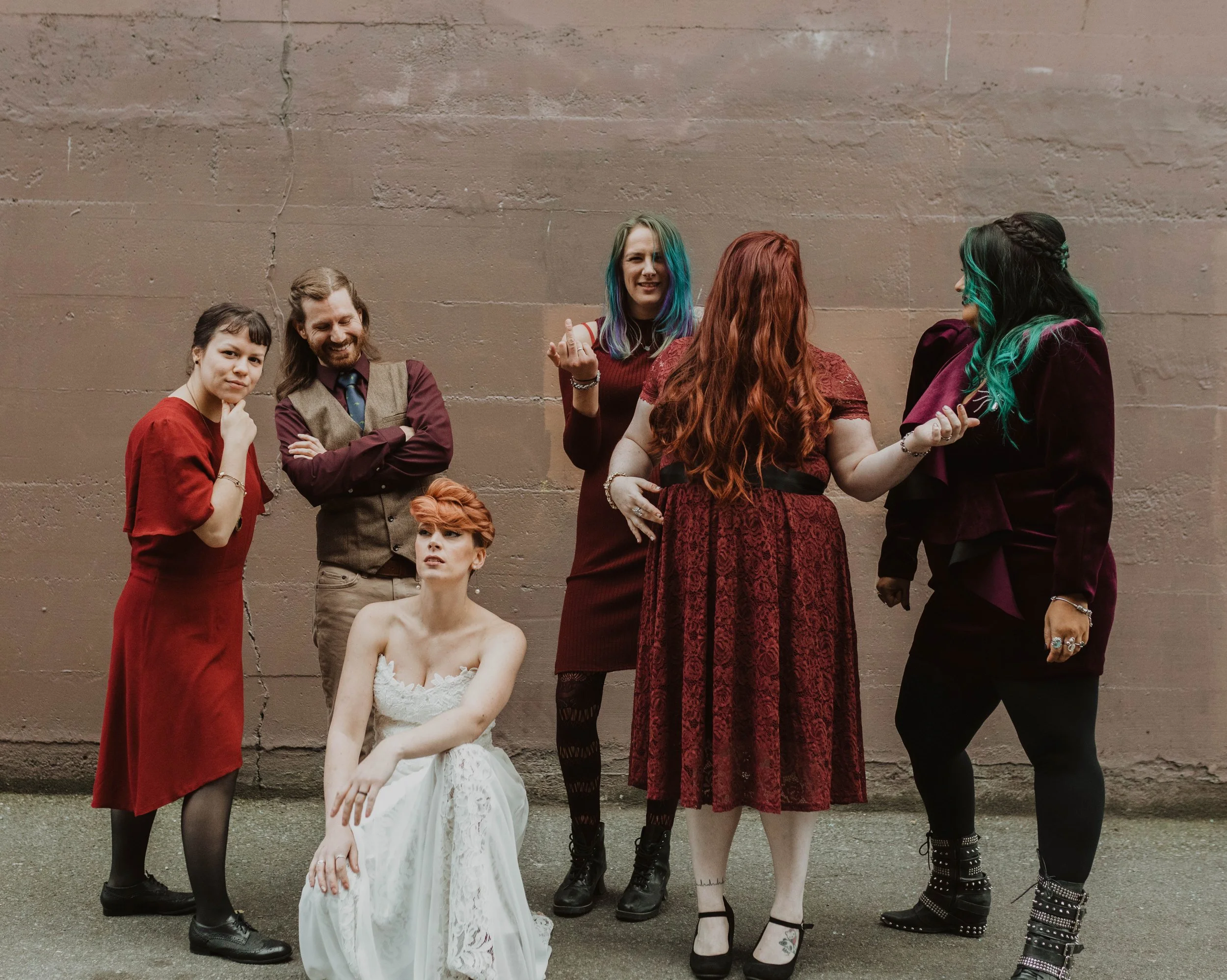 Six people, five women and one man, standing against a plain pink wall, with one woman sitting in front. They are dressed in various styles, some in red and burgundy outfits, and one woman with blue and green hair.