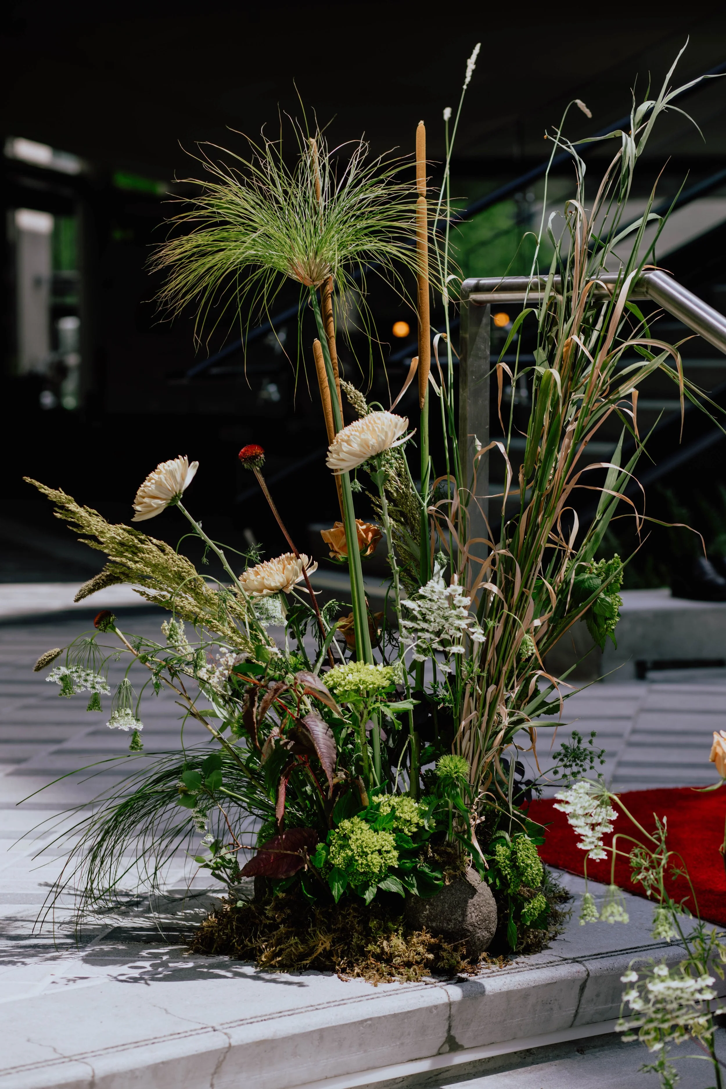 Arrangement of various flowers and plants including grasses, white and orange flowers, and moss at the base, placed on a tiered surface in a dark indoor setting. Seattle event photography