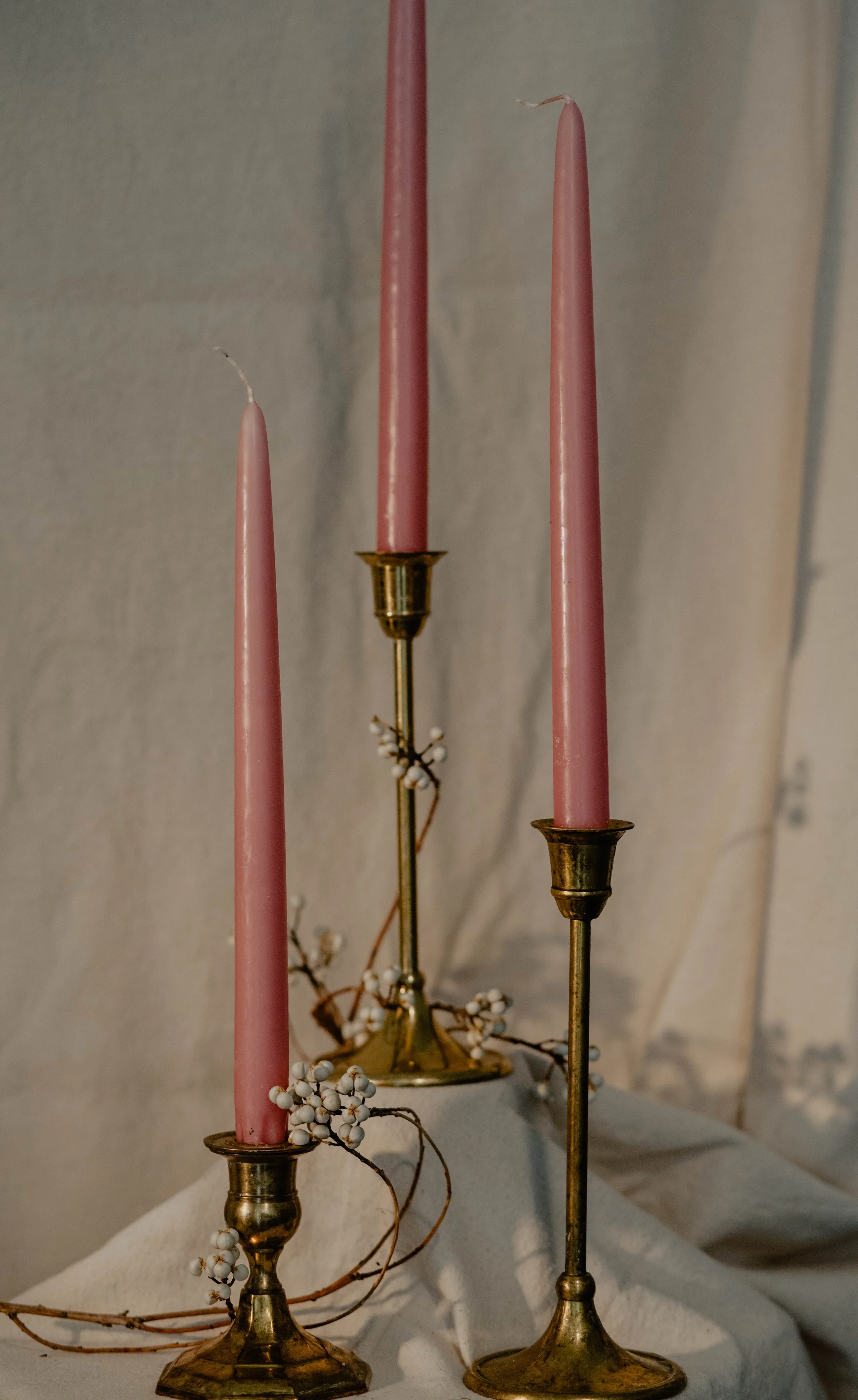 Three tall pink candles on vintage brass holders, with white berries and branches as decoration, set against light-colored curtains.  Seattle product photography