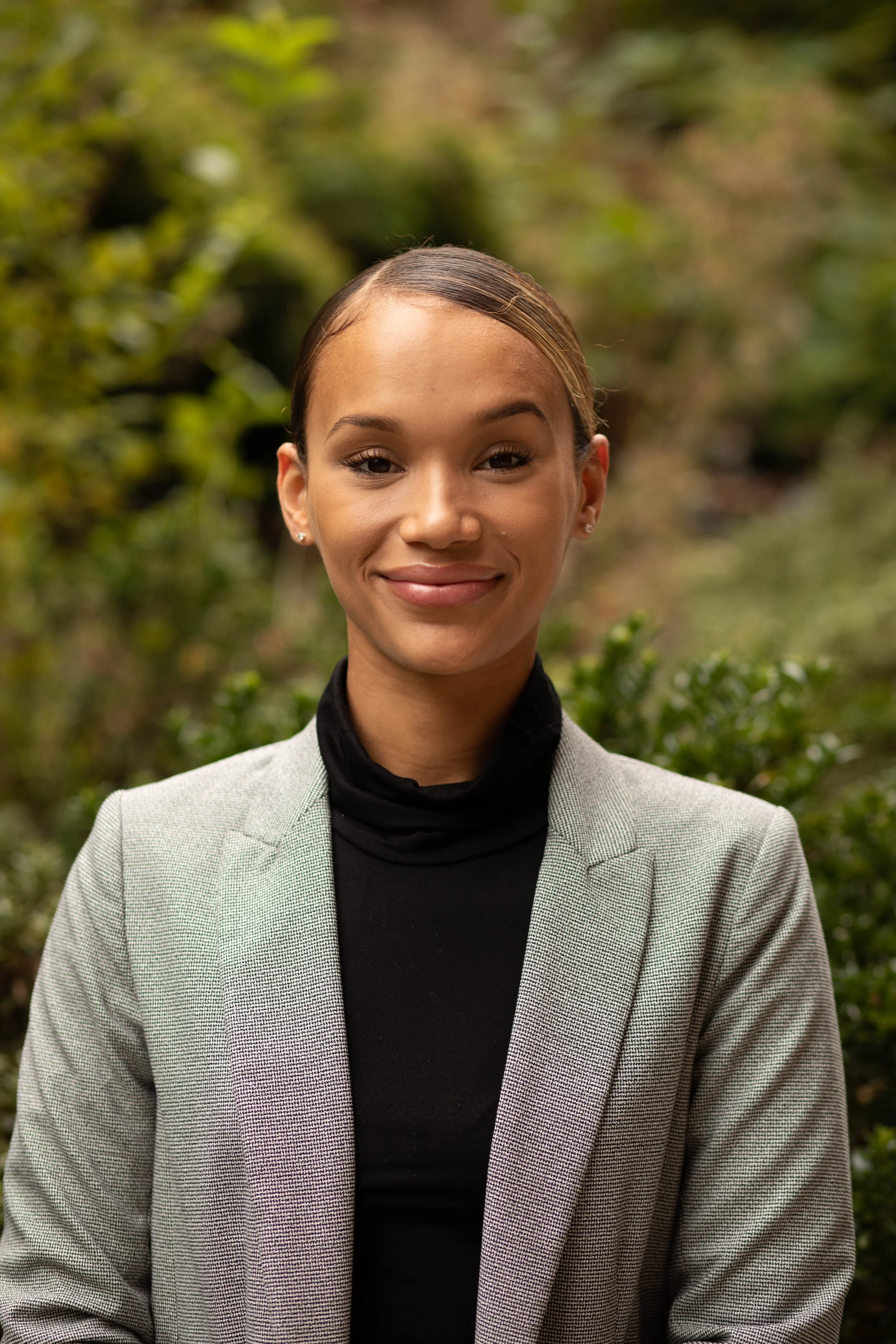 A young woman with light brown skin, wearing a grey blazer and a black turtleneck, standing outdoors in front of greenery with a friendly smile. Seattle professional head shot photography