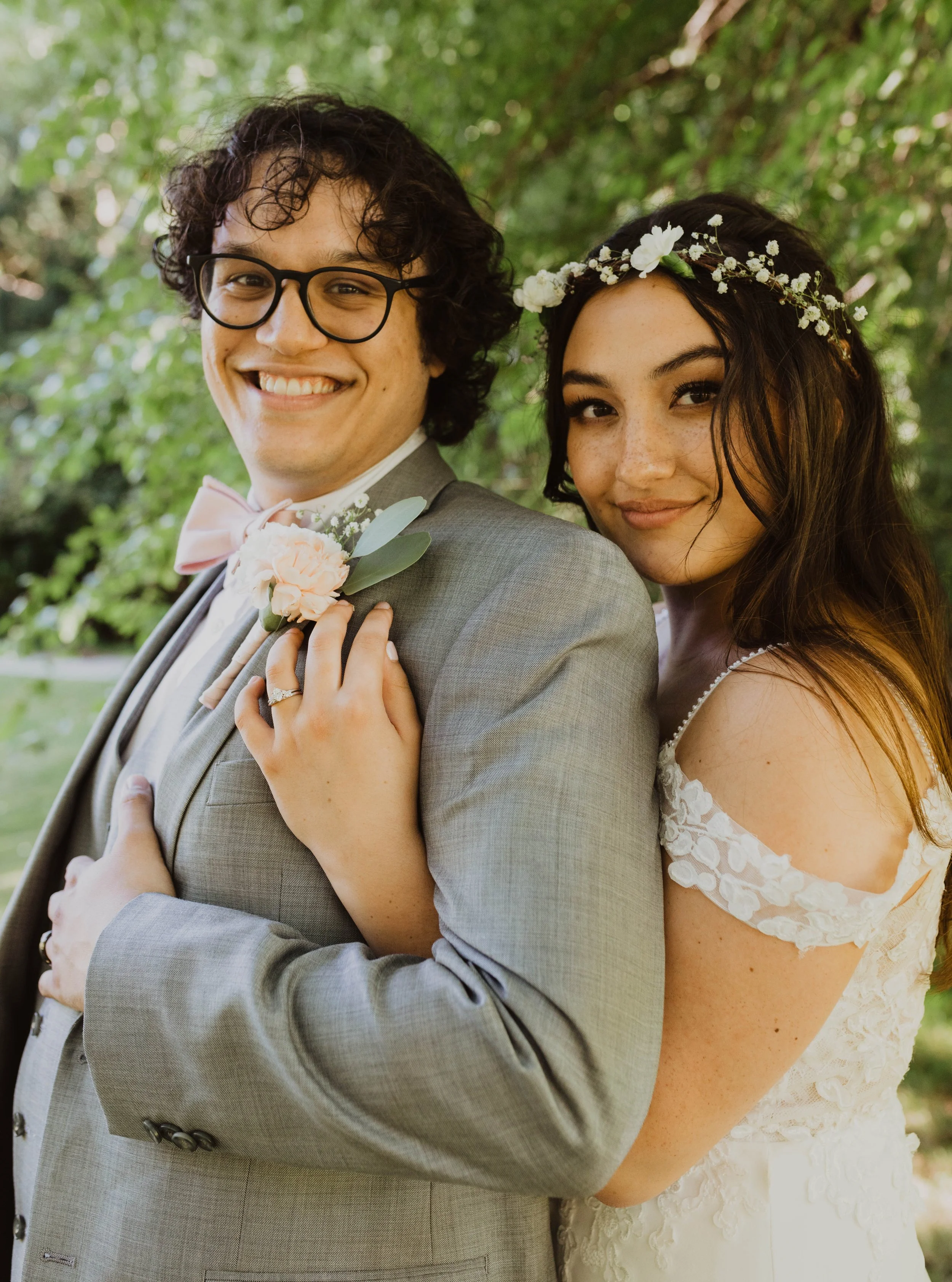 A smiling couple, the man wearing glasses and a light gray suit with a pink bow tie and boutonniere, and the woman in a white lace wedding dress with a flower crown, standing outdoors with greenery in the background. Wedding photography Seattle, WA.