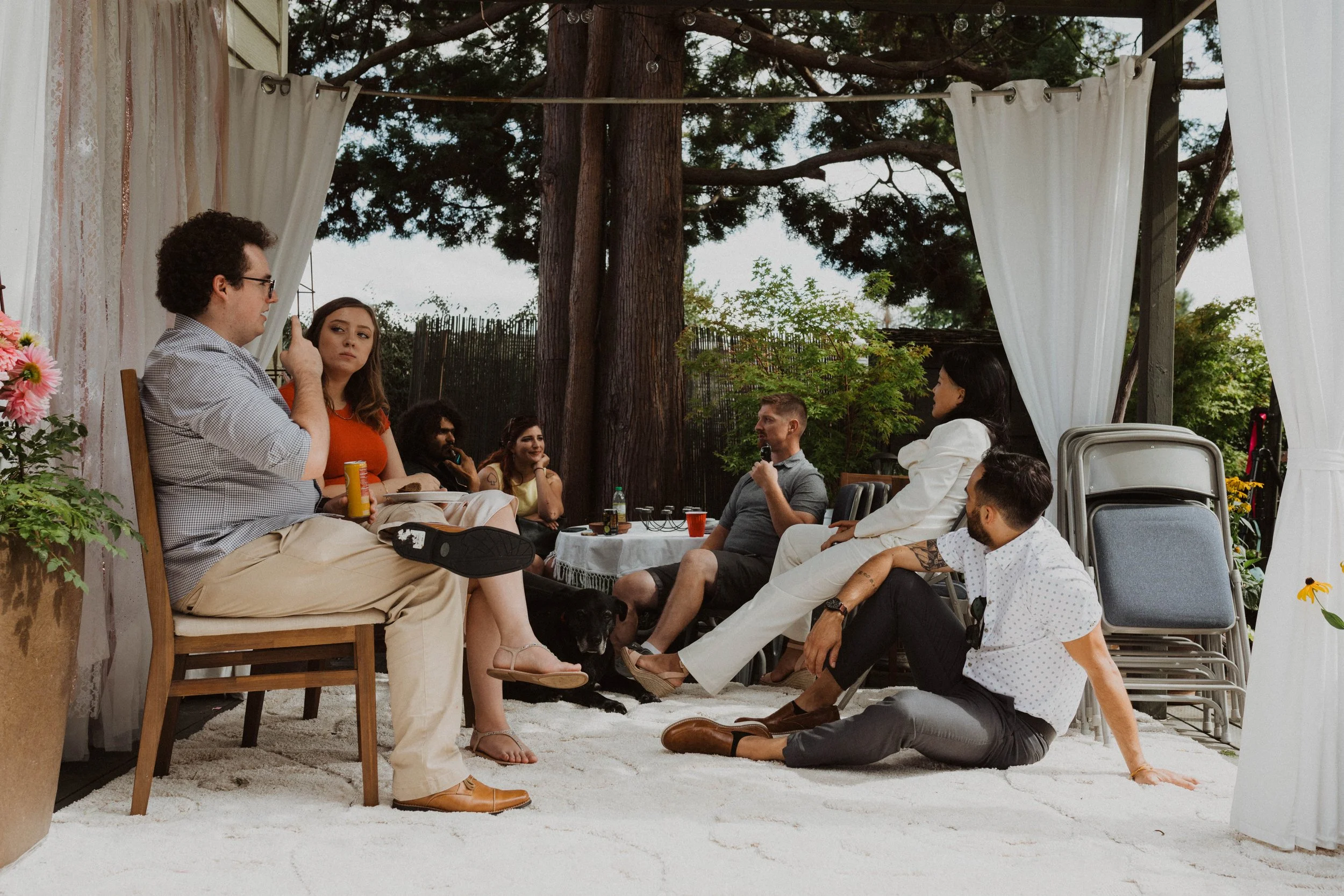 Group of people sitting and talking under a white canopy outdoors, with large trees and greenery in the background. Seattle, WA wedding photography.