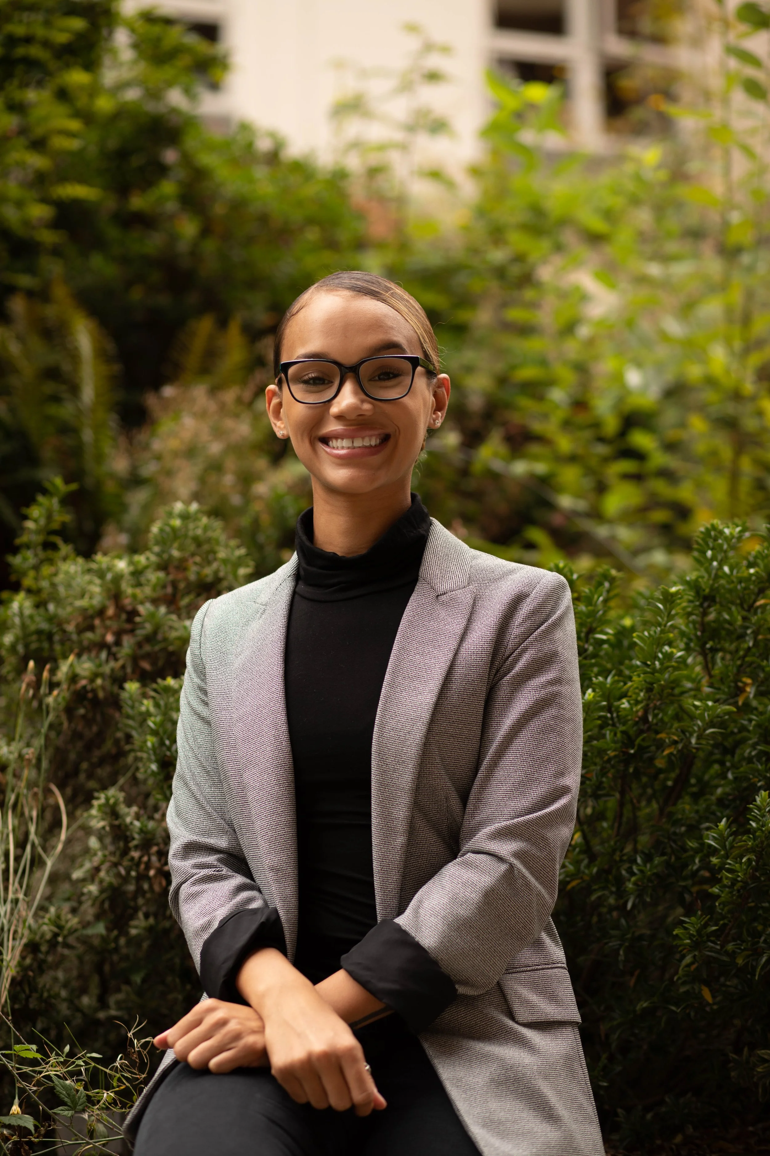 A woman with glasses, wearing a blazer and black turtleneck, sitting outdoors in front of green foliage, smiling at the camera. Seattle professional head shot photography