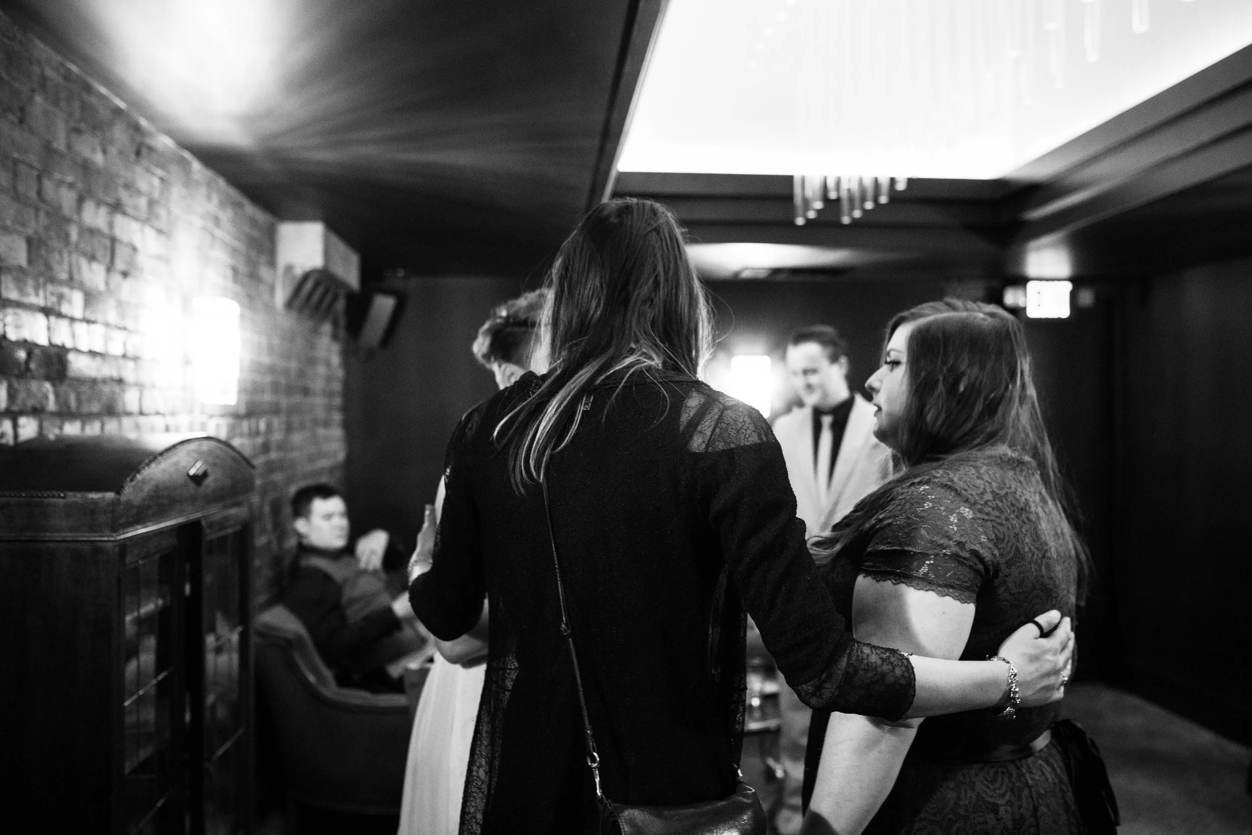 Black and white photo of four women in conversation in a dimly lit lounge, with a man seated in the background. Pioneer Square, Seattle, WA wedding photography.