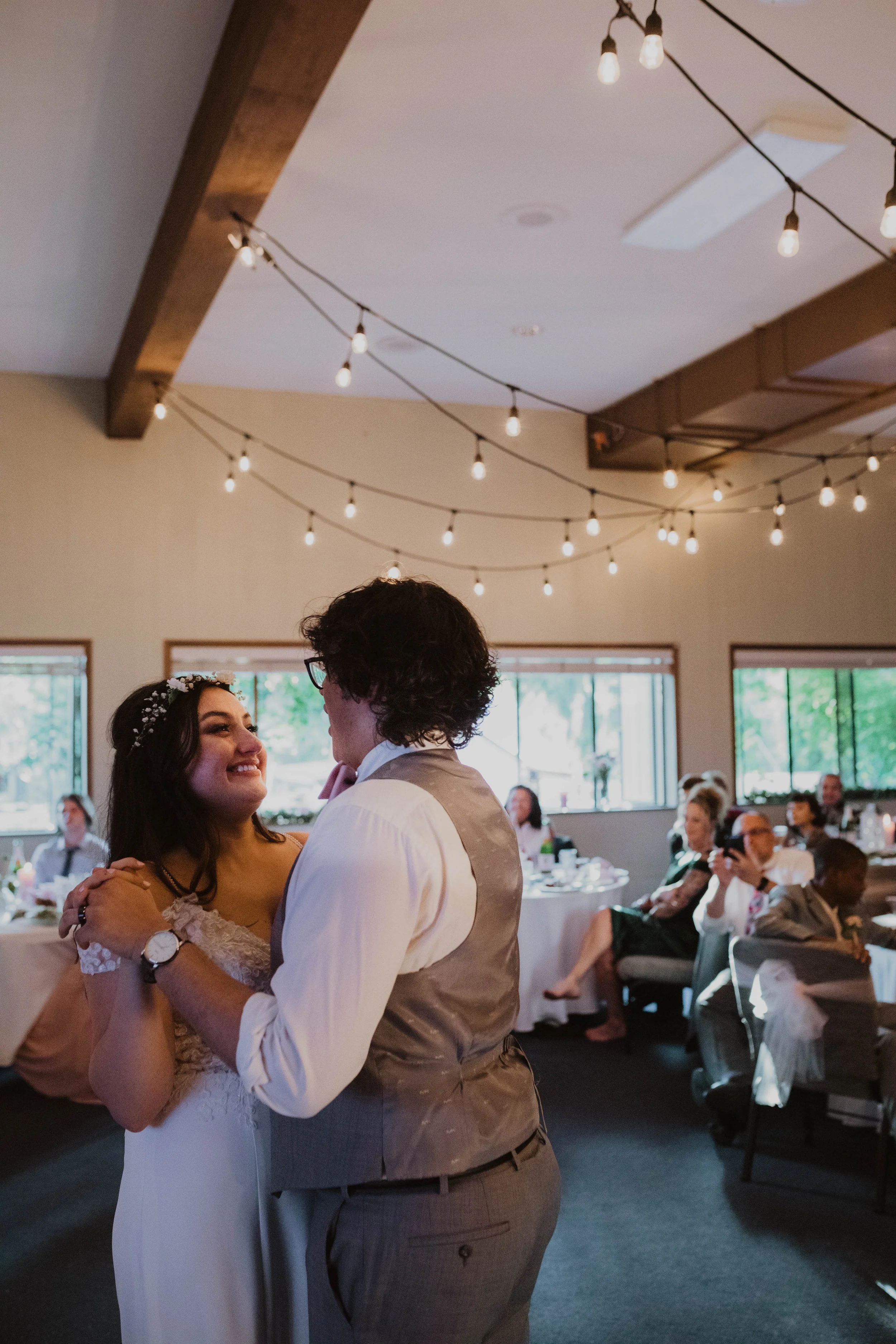 A couple dancing at a wedding reception with guests seated at tables in the background, decorated with string lights hanging from the ceiling. Seattle, WA wedding photography.