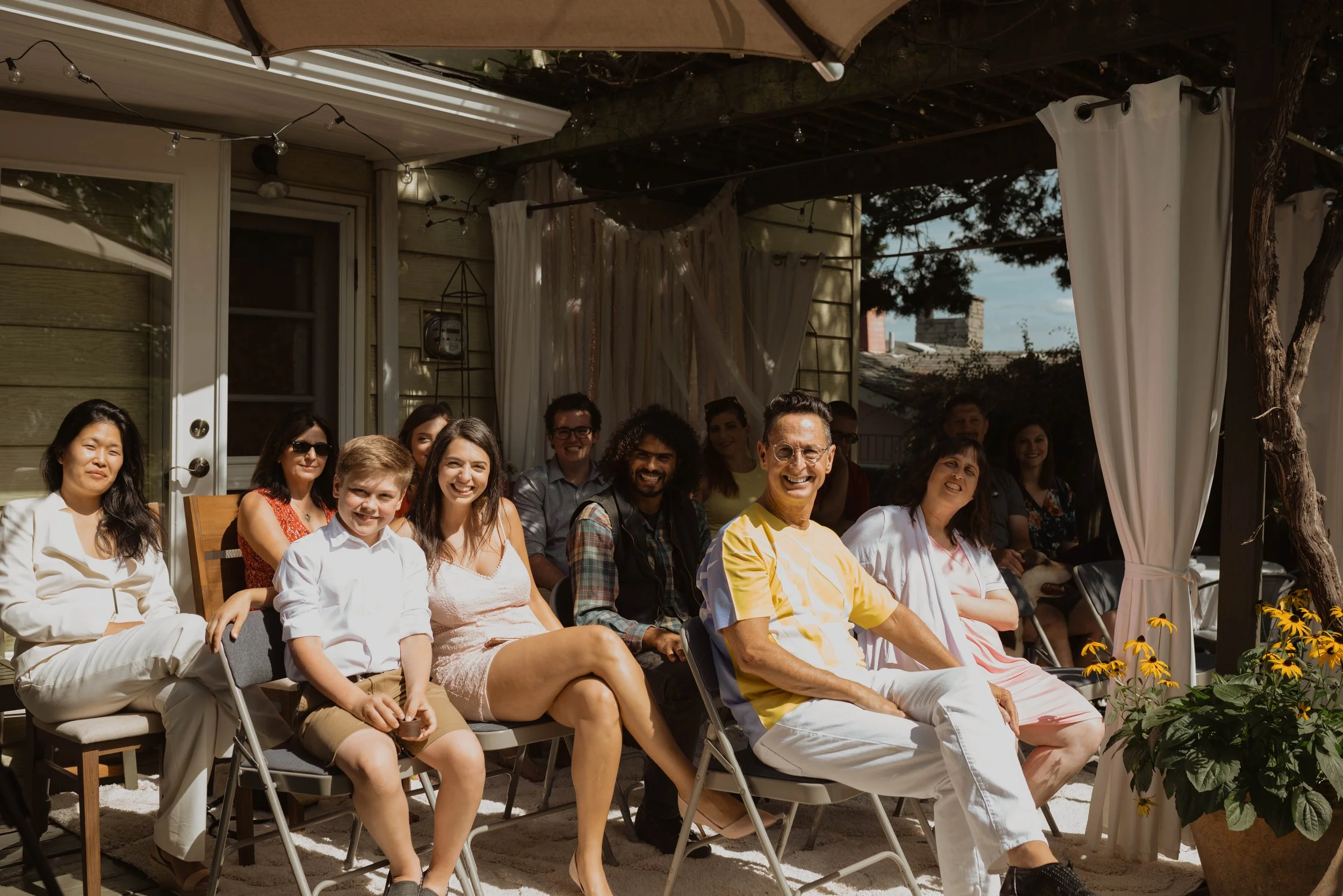Group of people enjoying an outdoor gathering on a patio, sitting on chairs, smiling, with a house and trees in the background. Seattle, WA wedding photography.