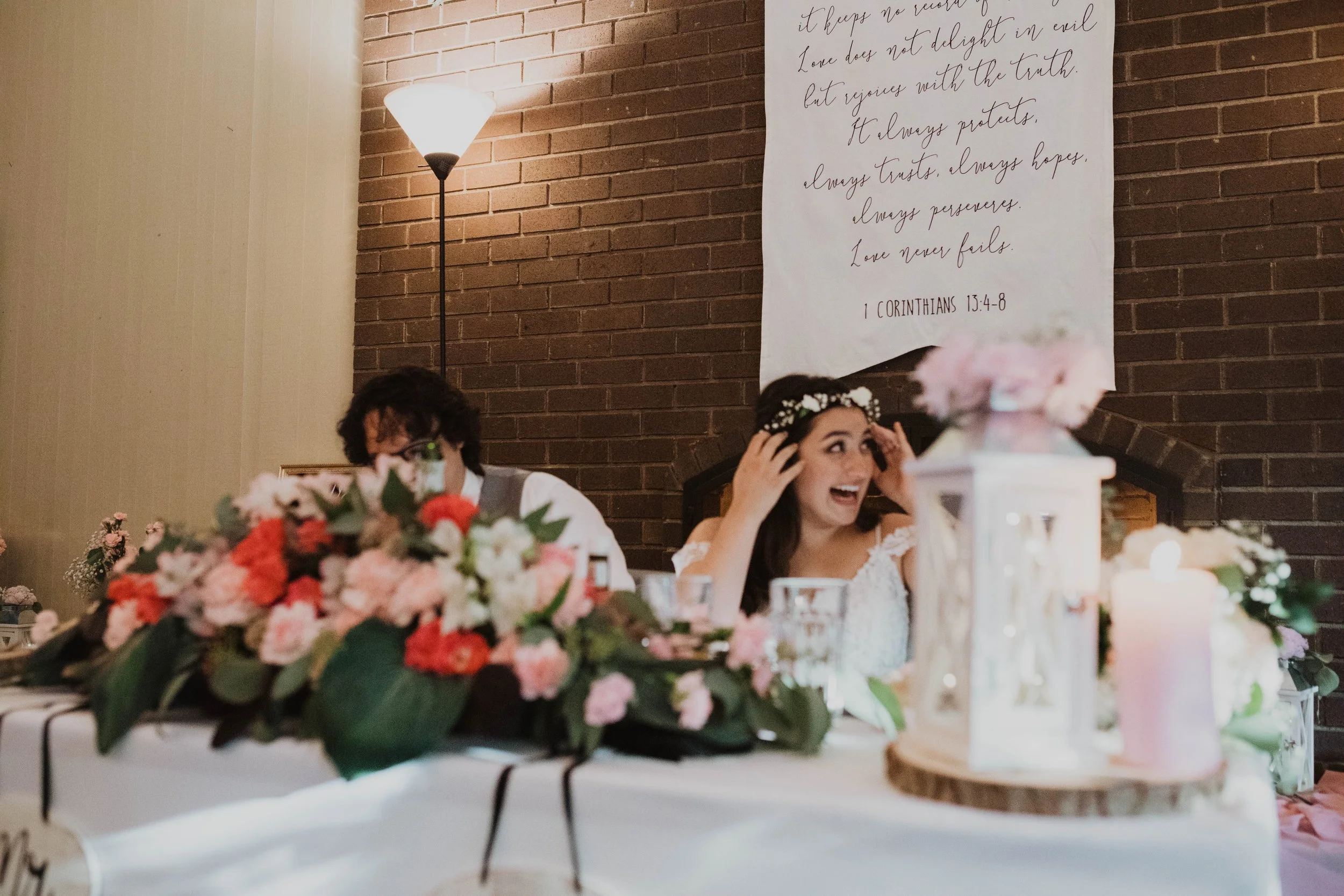 A bride and a man sitting at a decorated wedding table, with a floral centerpiece and candles, smiling and laughing. A large religious quote is on the brick wall behind them. Seattle, WA wedding photography.