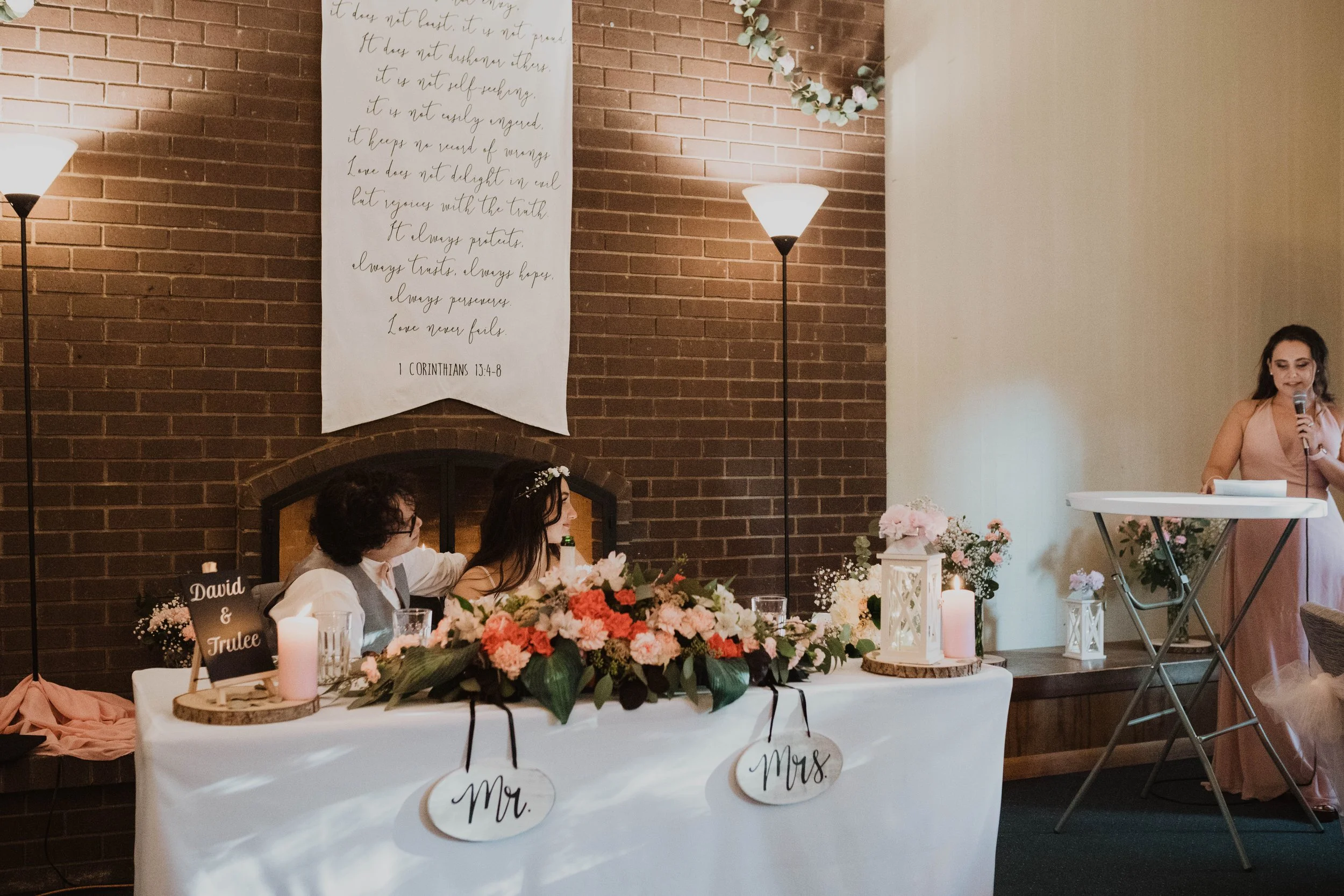 A wedding ceremony with a bride and groom seated at a table decorated with flowers, candles, and signs reading 'Mr.' and 'Mrs.' in front of a brick wall. A woman in pink dress stands at a lectern, reading from a paper, speaking into a microphone in a