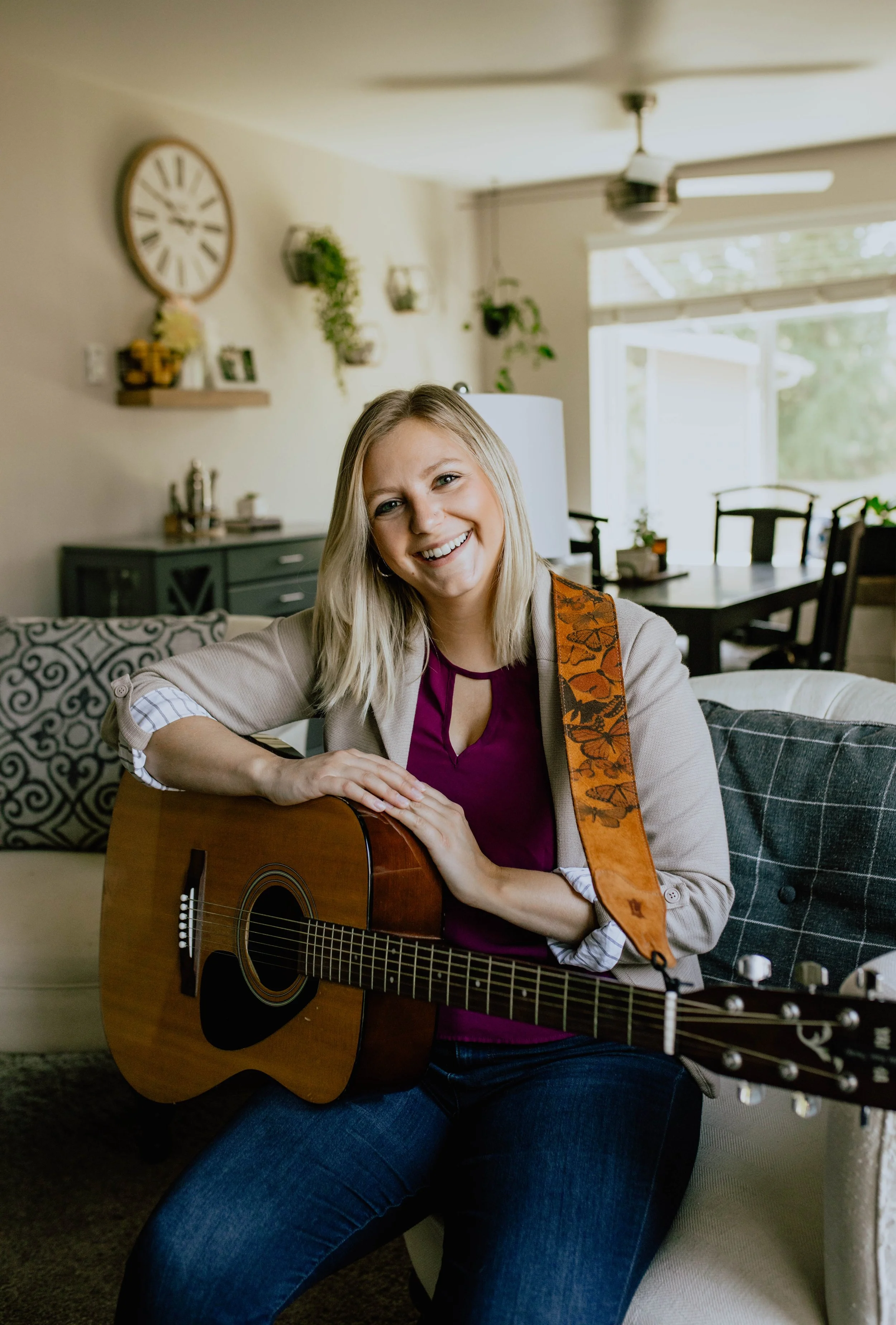 A woman with blonde hair smiling, sitting on a couch in a living room, holding an acoustic guitar. Seattle professional head shot photography