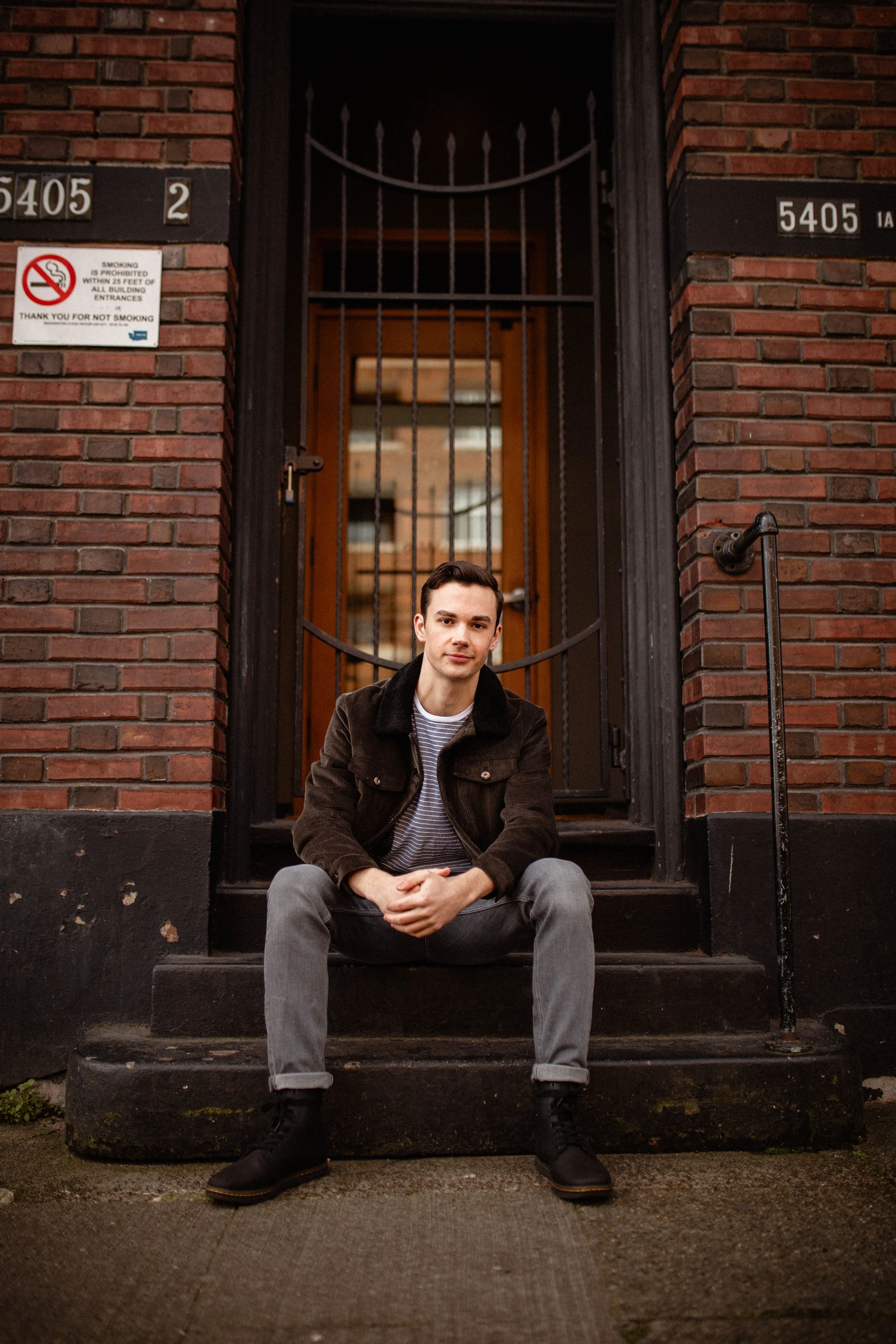 A young man in casual clothing sitting on the steps of a brick building, with a metal gate and building windows behind him. Seattle professional head shot photography