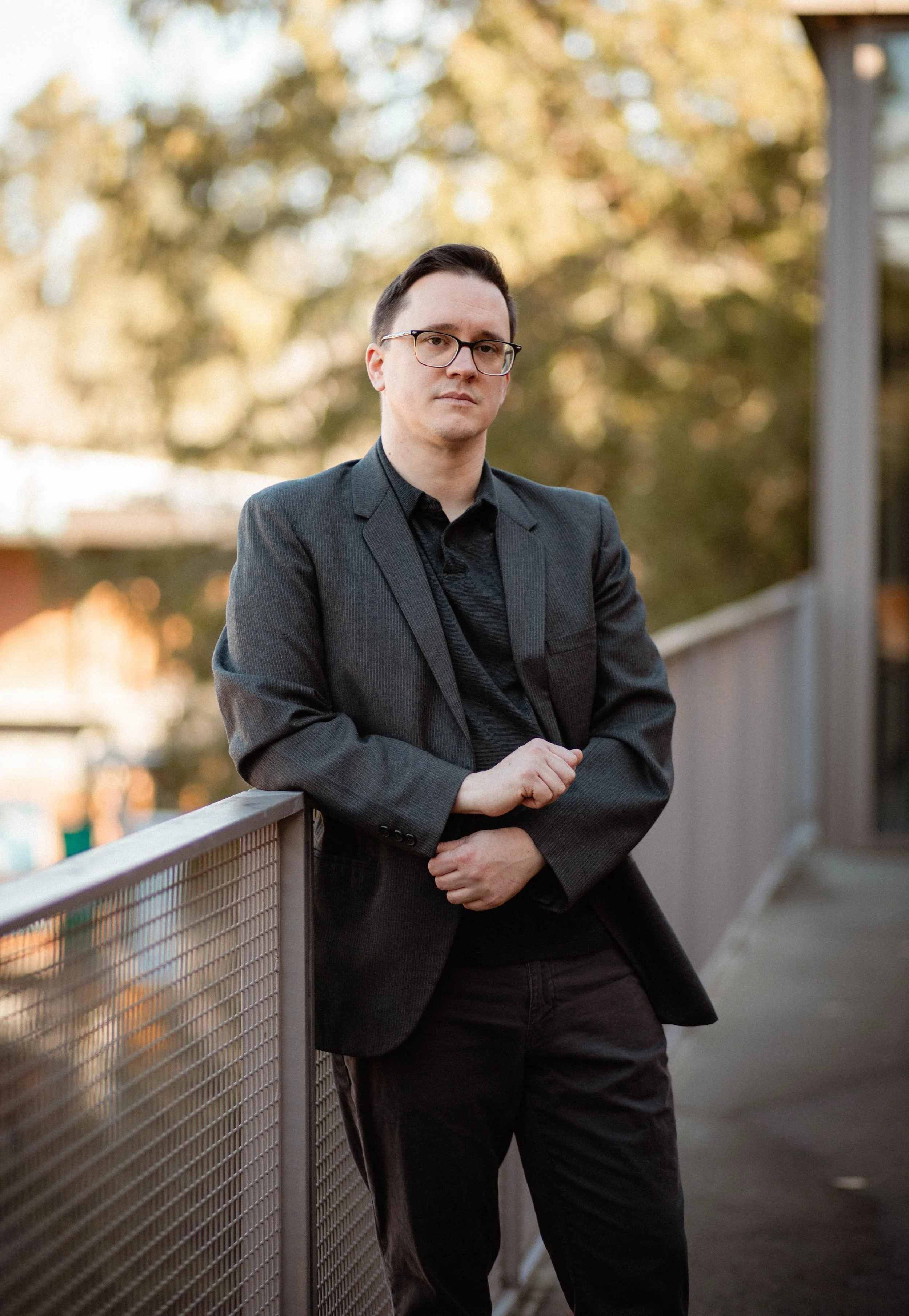 A man in a black blazer and glasses standing outdoors leaning against a railing with blurred trees in the background. Seattle professional head shot photography