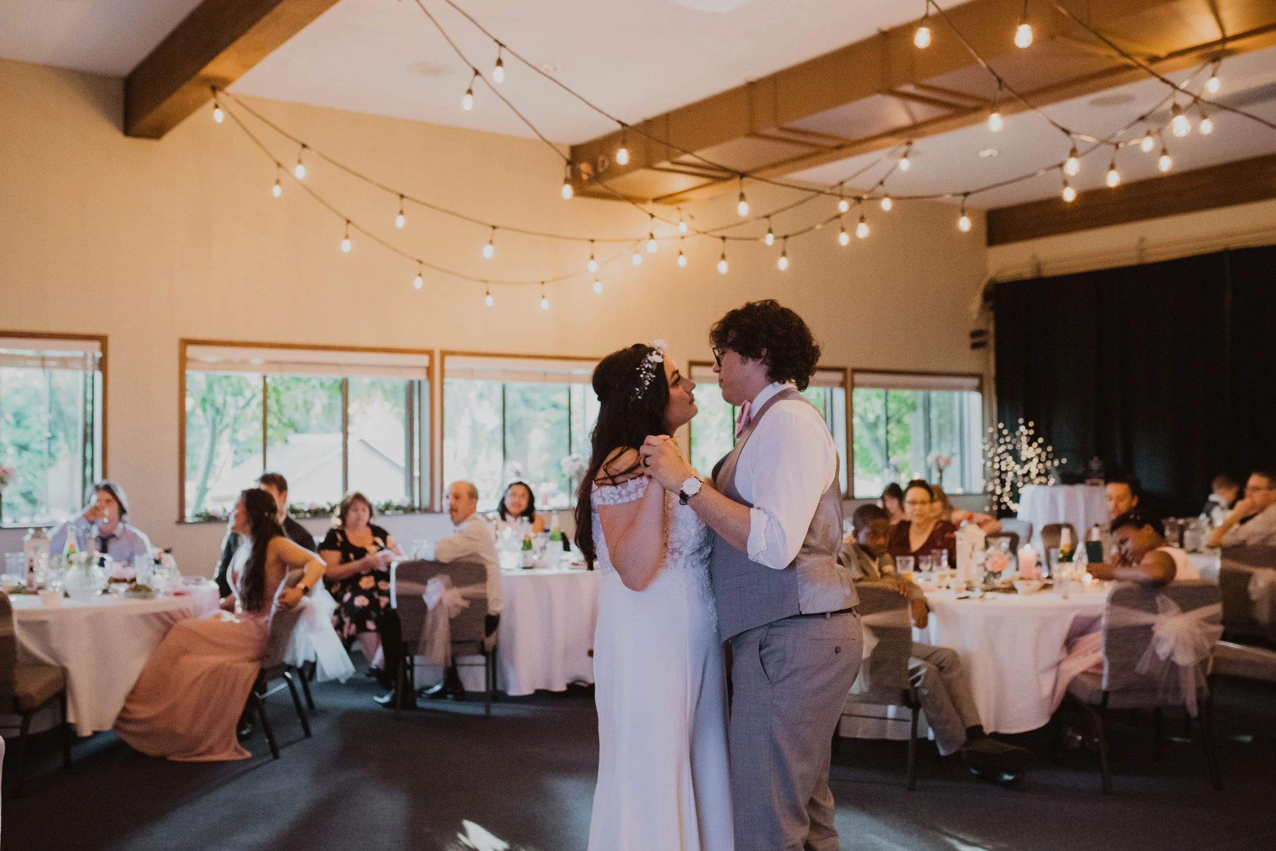 A bride and groom share a dance at their wedding reception in a decorated banquet hall with string lights, with seated guests watching in the background. Seattle, WA wedding photography.