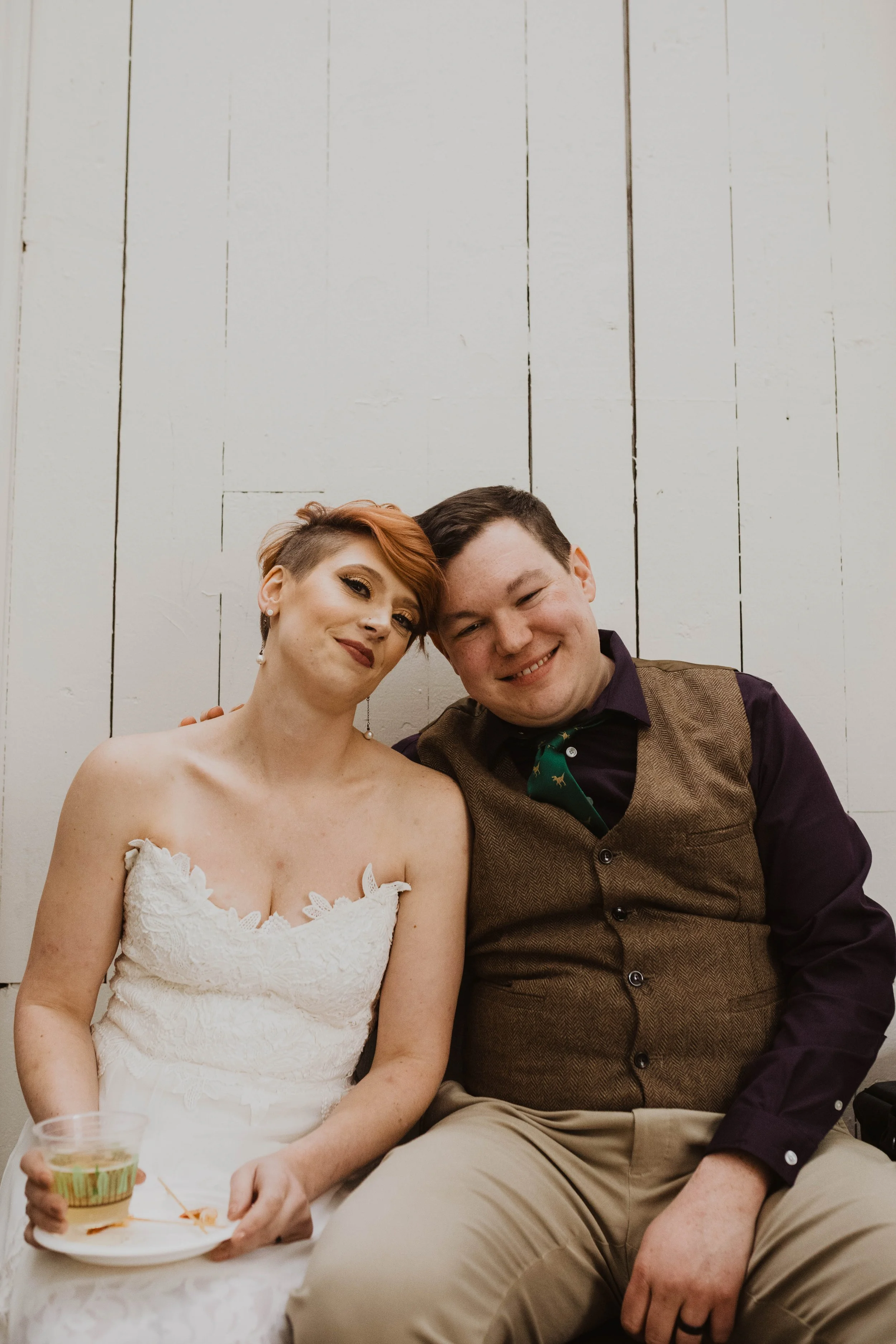 A couple sitting close together, smiling at the camera against a white wooden wall, the woman in a white lace dress holding a drink and a plate, and the man in a brown vest and dark shirt. Pioneer Square, Seattle, WA wedding photography.
