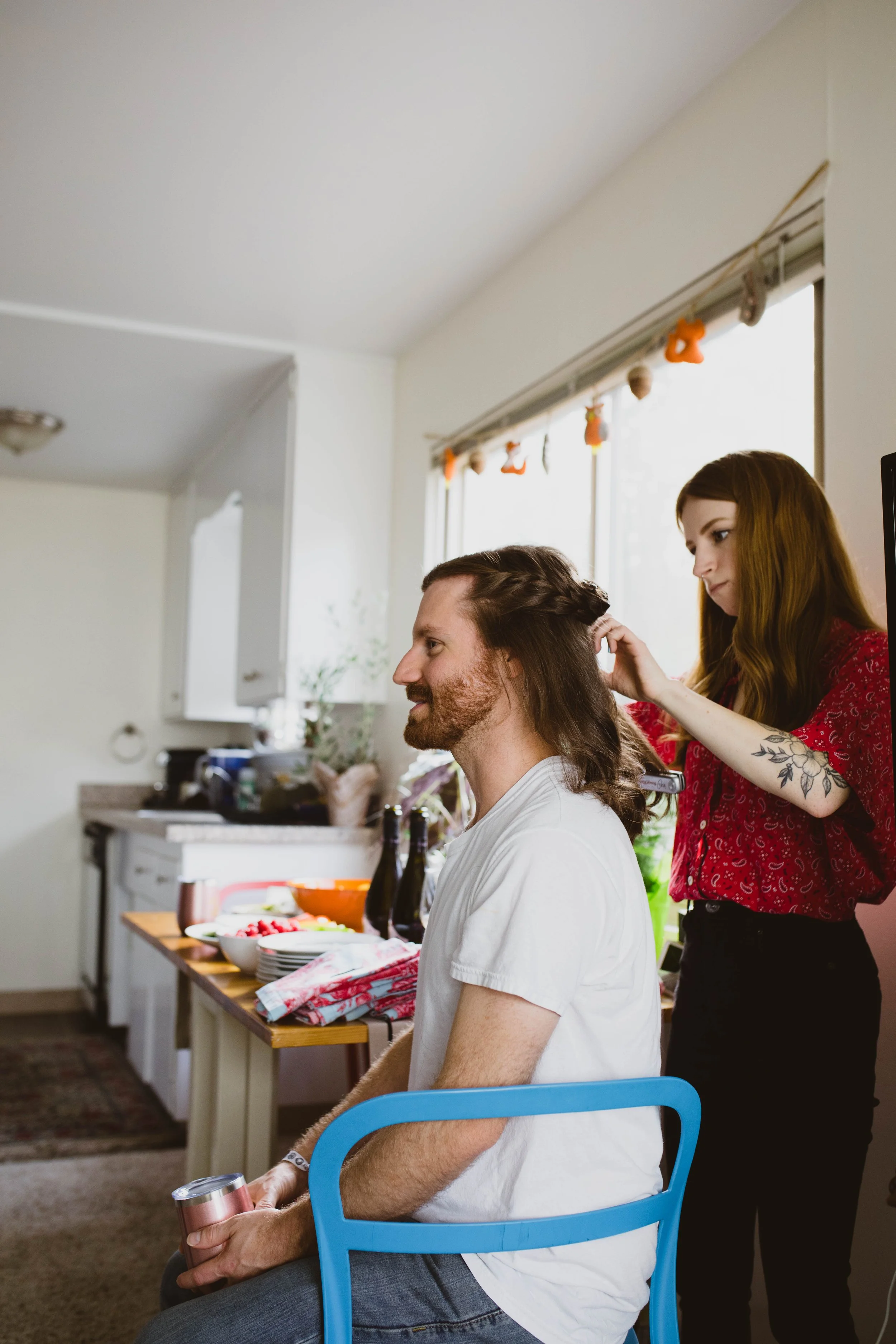 A woman with long brown hair, tattoos on her arm, wearing a red blouse, is styling a man's long brown hair in a kitchen with white cabinets and a window with hanging decorations, while he is sitting on a blue chair holding a canned drink.