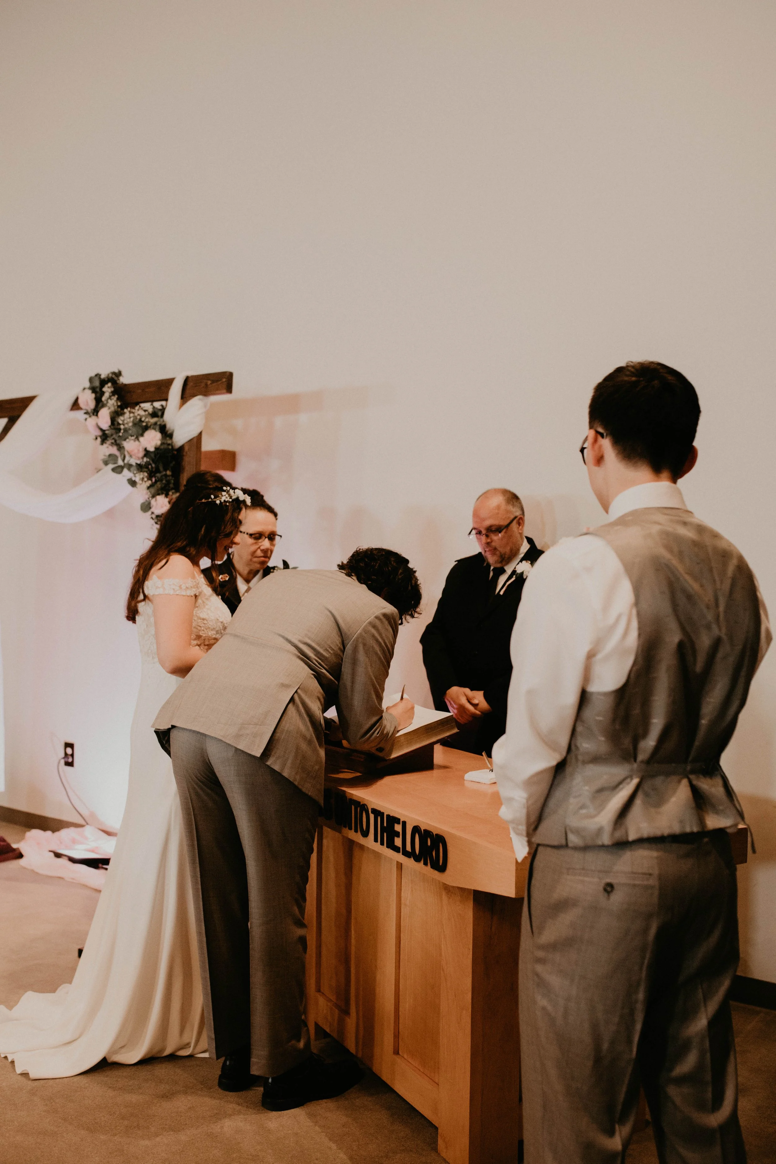 A wedding ceremony with the bride and groom signing a marriage document, surrounded by witnesses and officiants, with a floral arch in the background. Seattle, WA wedding photography.