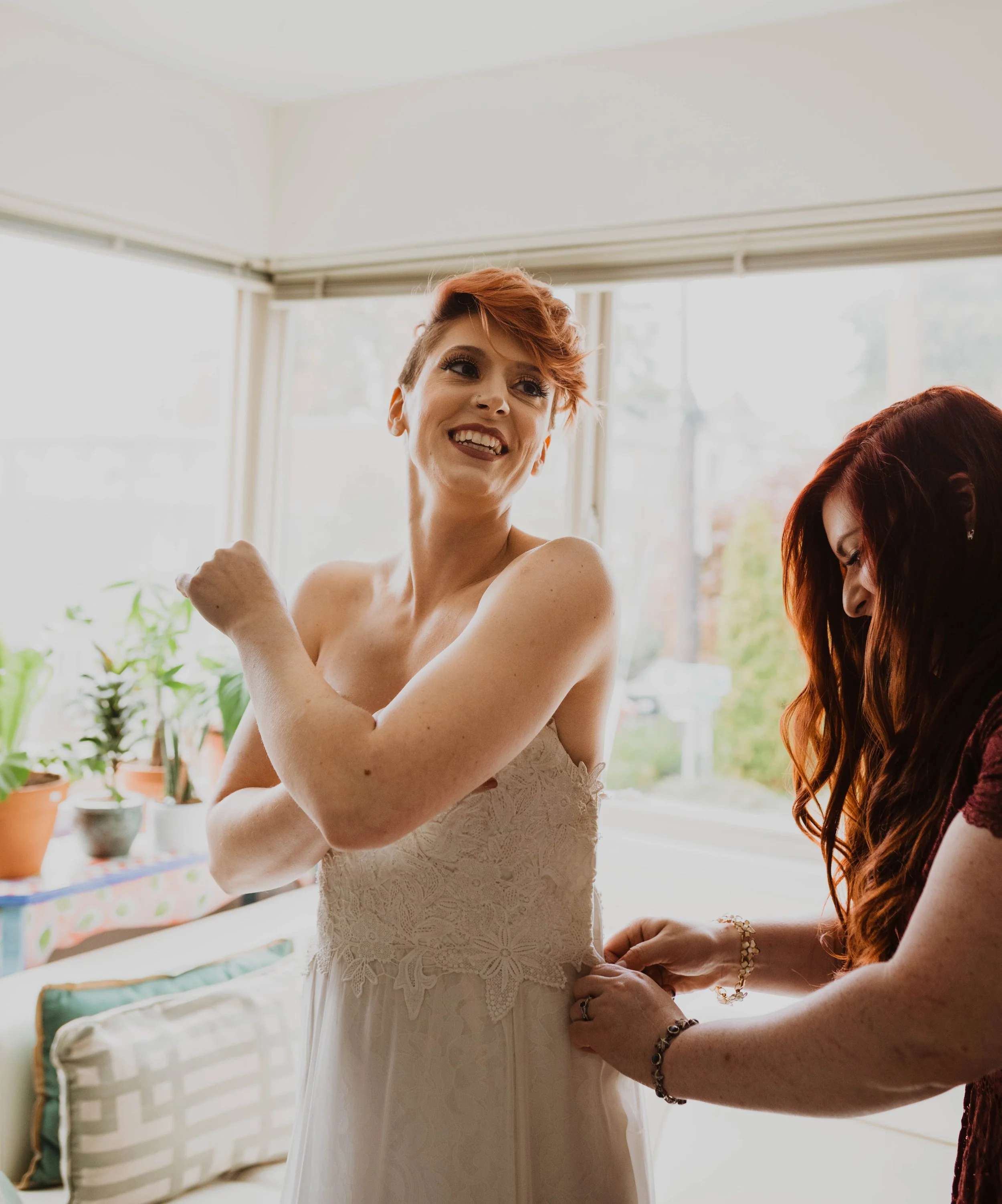 A bride with short red hair is smiling and adjusting her dress while another woman with long red hair helps her. Pioneer Square, Seattle, WA wedding photography.