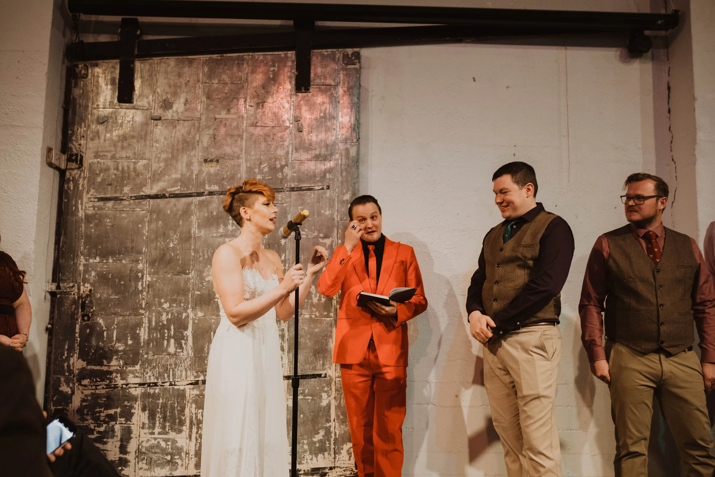 Group of four people standing on stage, one woman in a white dress speaking into a microphone and three men dressed in business casual attire listening. Pioneer Square, Seattle, WA wedding photography.