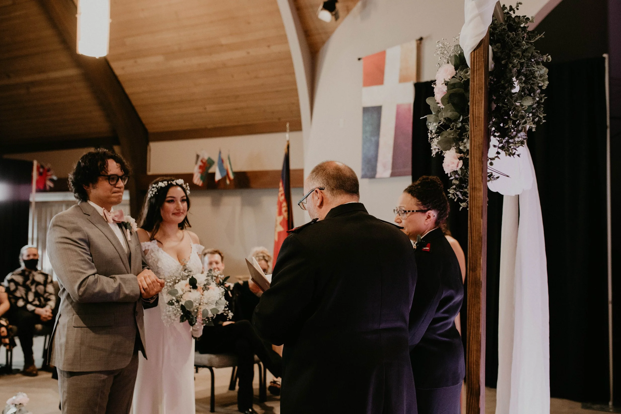 A wedding ceremony with a bride in a white dress holding a bouquet, a groom in a light-colored suit, and two officiants in black suits. The background features flags and artistic decorations, with guests seated and watching.