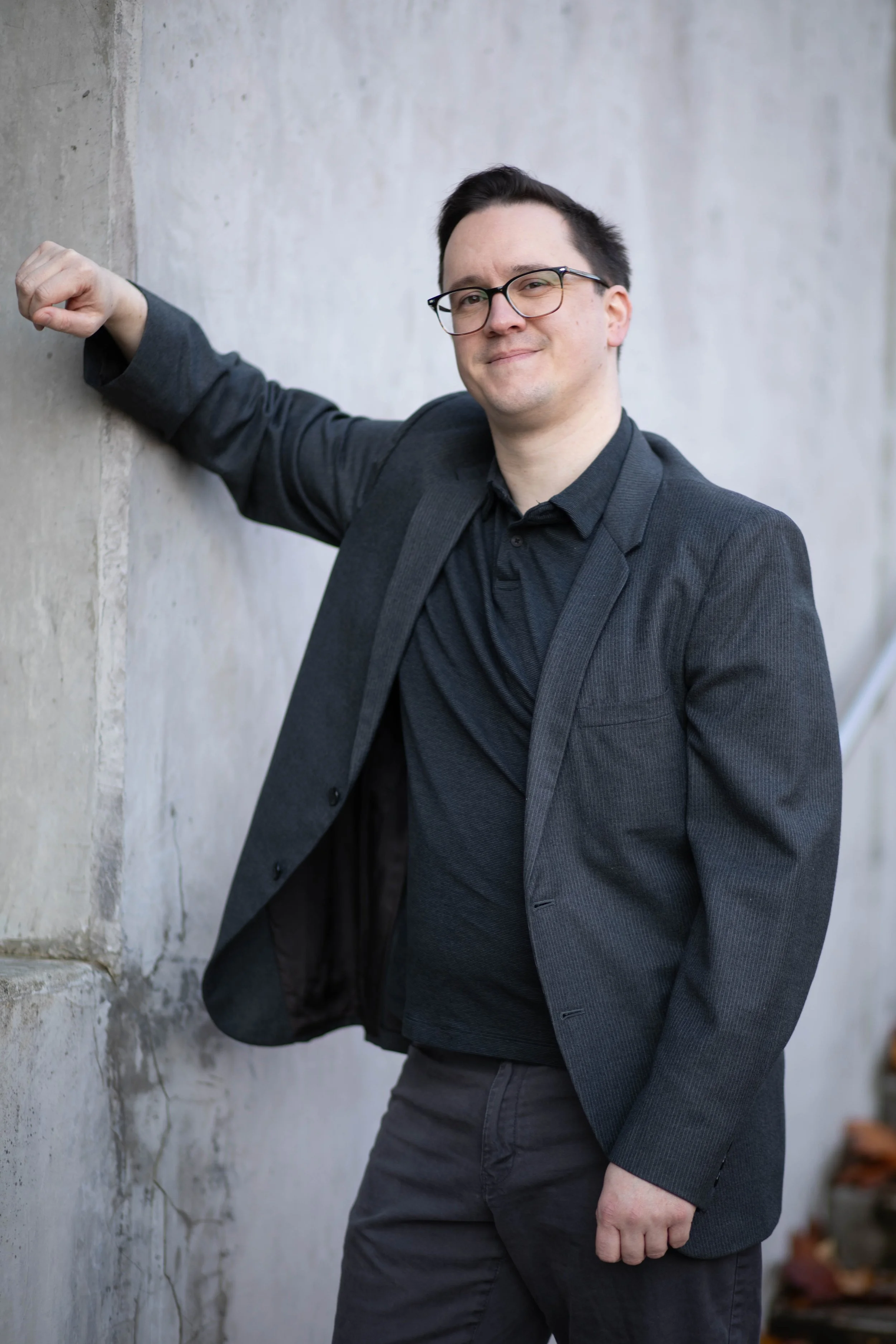 A man wearing glasses in a black shirt and dark gray blazer leaning against a concrete wall with one arm extended and smiling. Seattle professional head shot photography