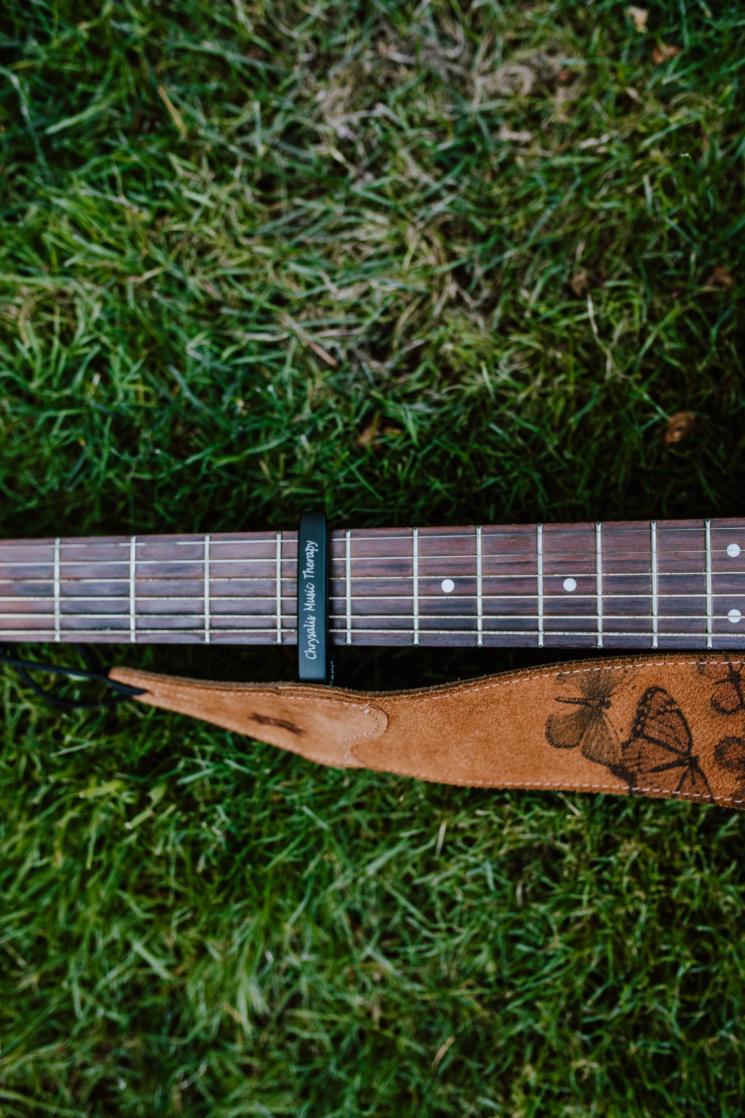 A guitar lying on grass with a brown strap and a black capo labeled 'Chopstick Music Therapy' on the fretboard. Seattle professional head shot photography