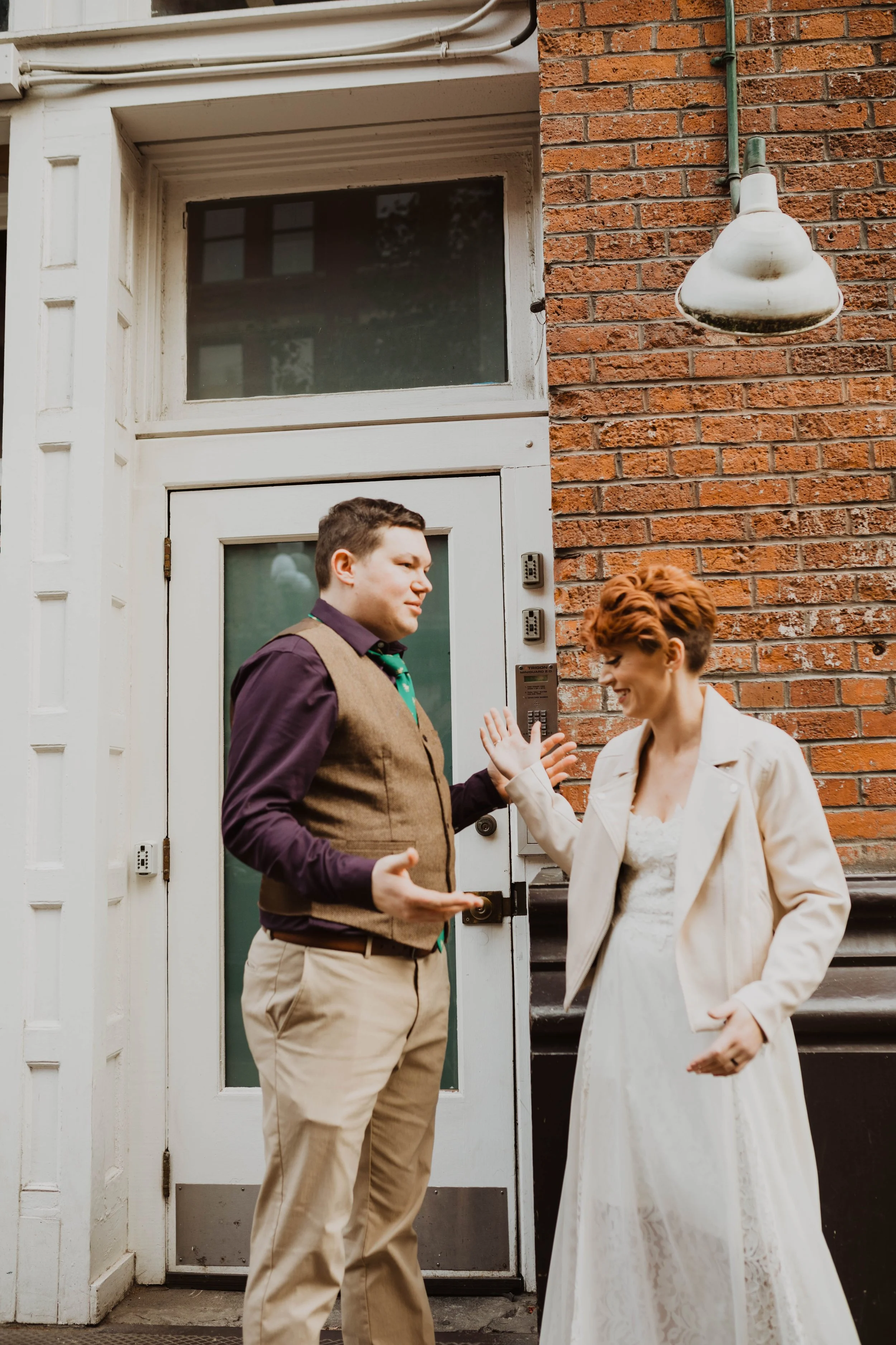 A man and a woman having a friendly conversation outdoors near a white door with a glass window, in front of a brick wall, during daytime. Pioneer Square, Seattle, WA wedding photography.