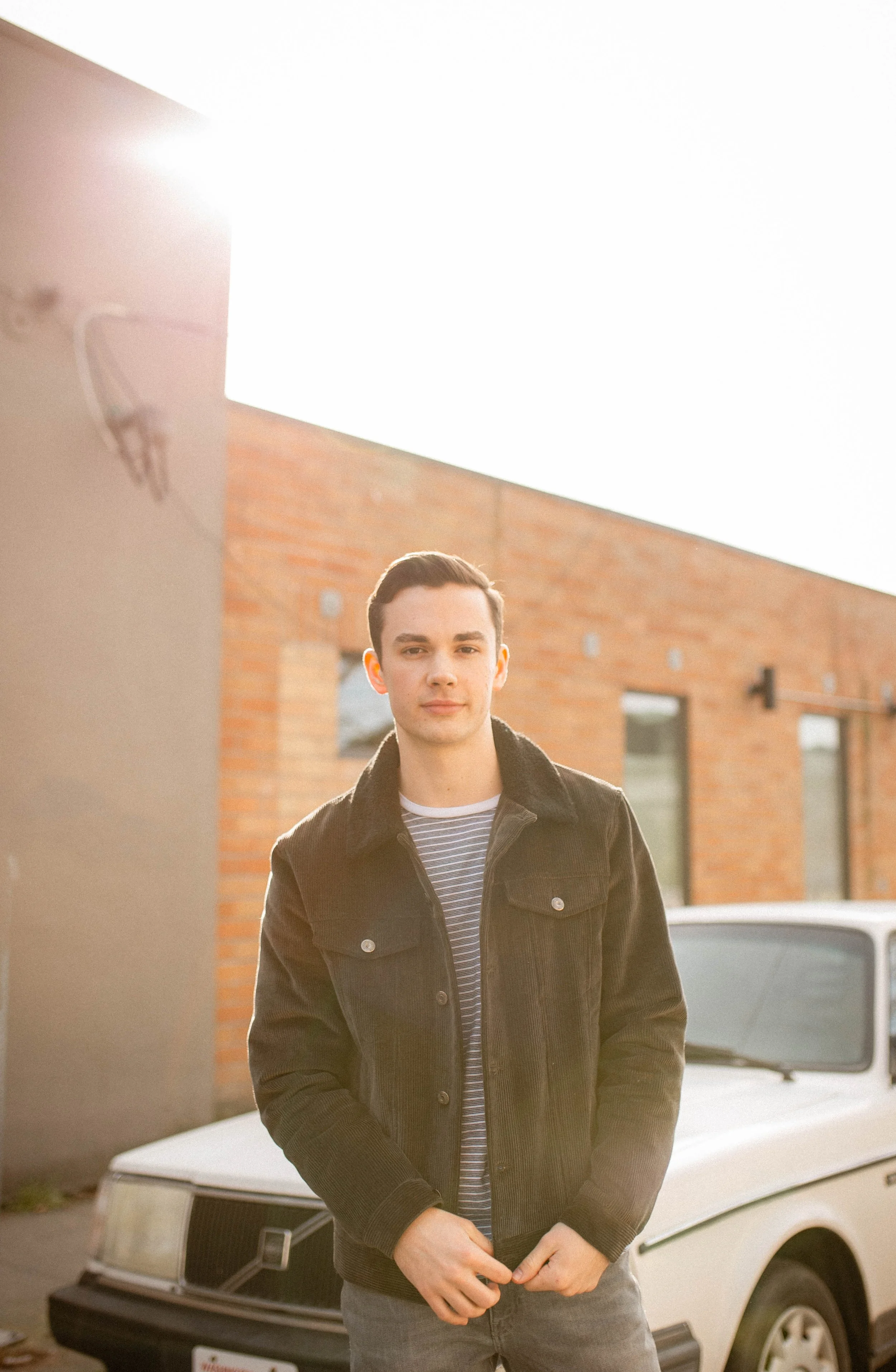 A young man in a black jacket and striped shirt standing outdoors near a vintage white car, with a brick building in the background, illuminated by warm sunlight. Seattle professional head shot photography