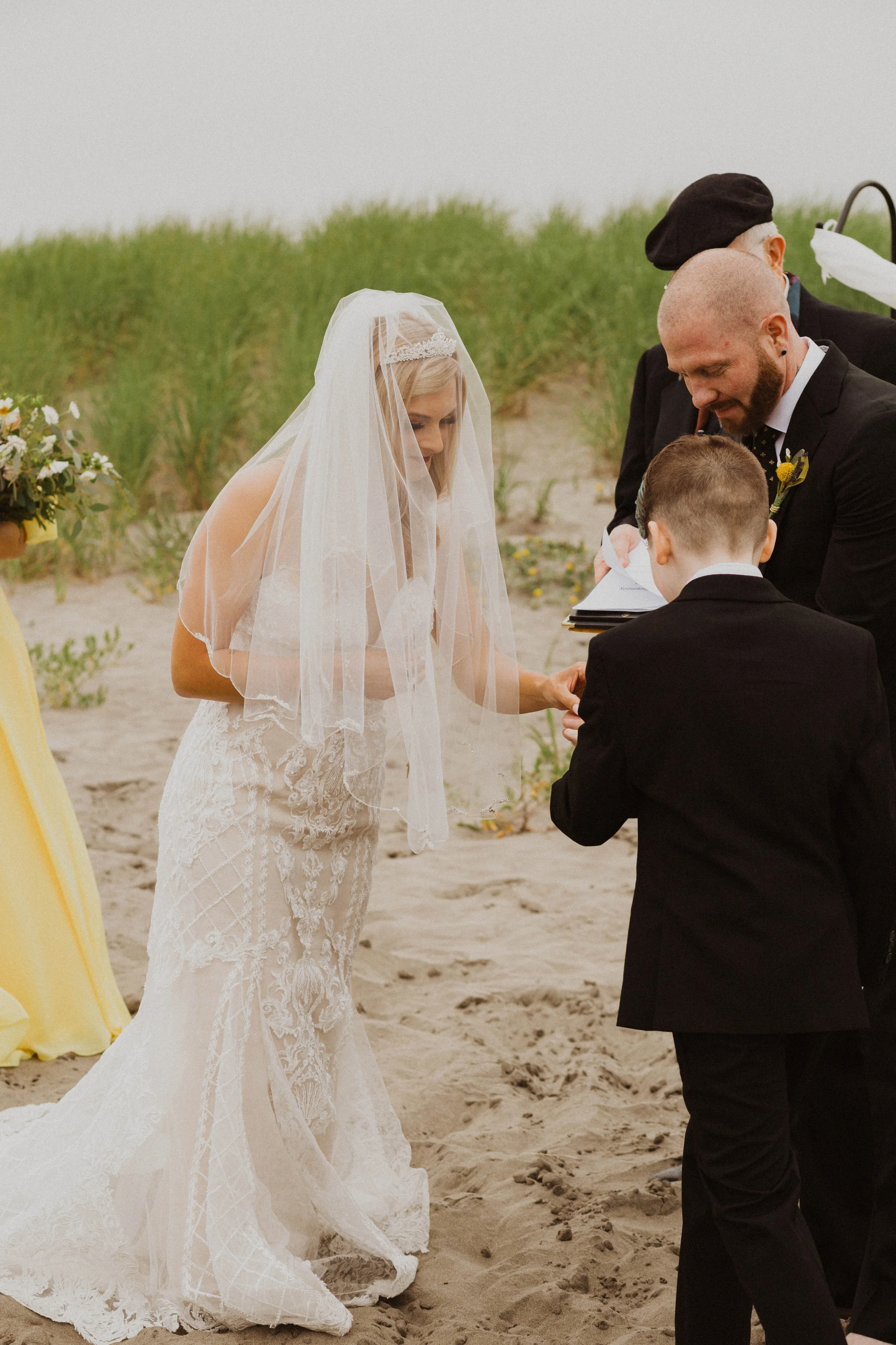 A beach wedding ceremony with a bride in a lace wedding dress and veil, a groom in a black suit, and a young boy in a tuxedo, with officiants present, on the sandy shore with green dunes in the background. Long Beach, WA wedding photography.