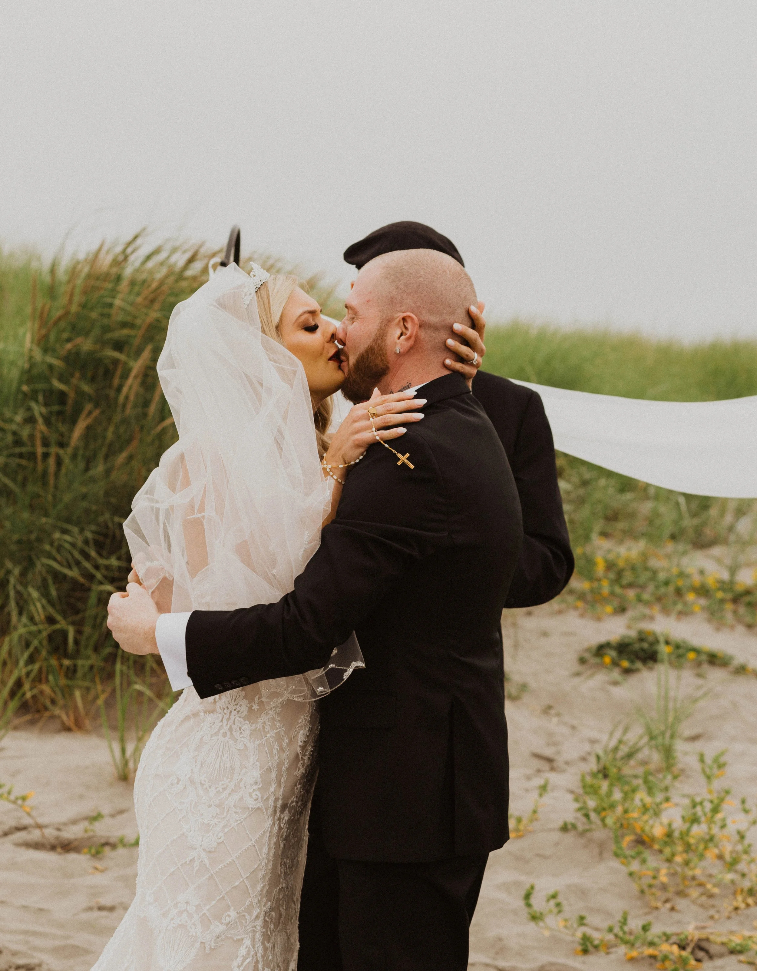 A bride and groom share a kiss at an outdoor beach wedding, with the bride wearing a lace wedding dress and veil and the groom in a black suit. Long Beach, WA wedding photography.