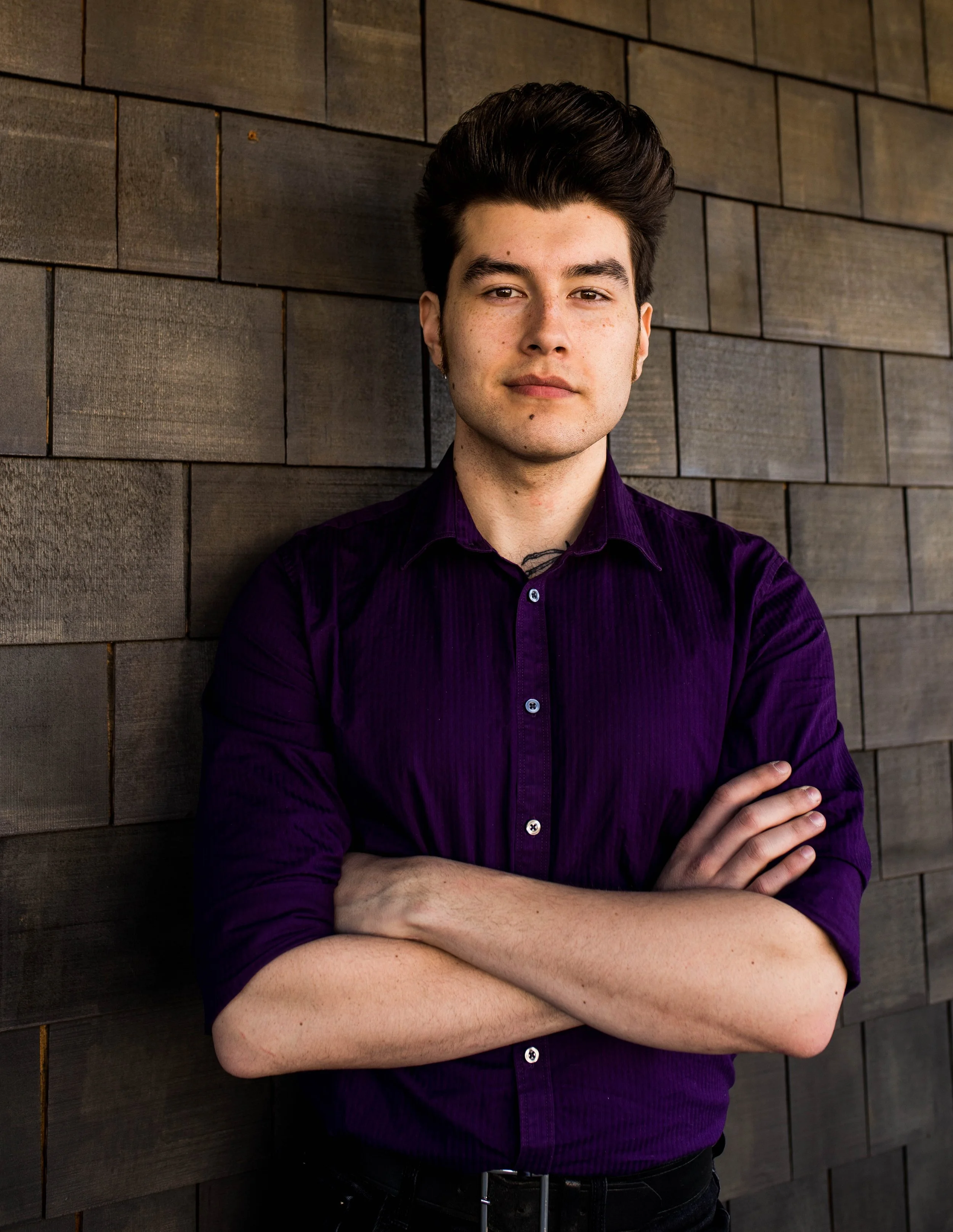A young man with dark hair, wearing a purple shirt, standing with arms crossed in front of a wooden wall. Seattle professional head shot photography