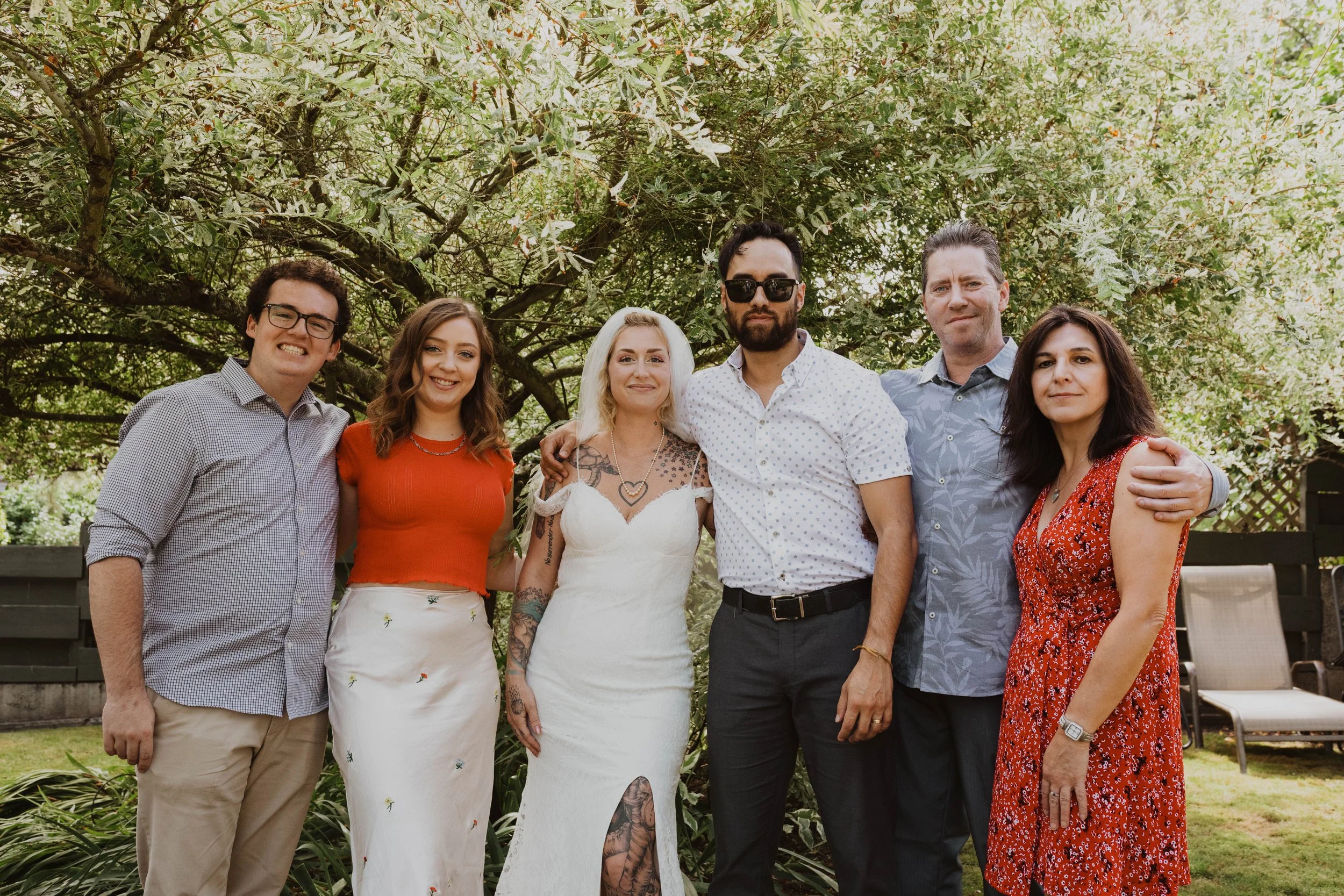 Group of six people smiling and posing outside under a large tree in a backyard. Seattle, WA wedding photography.