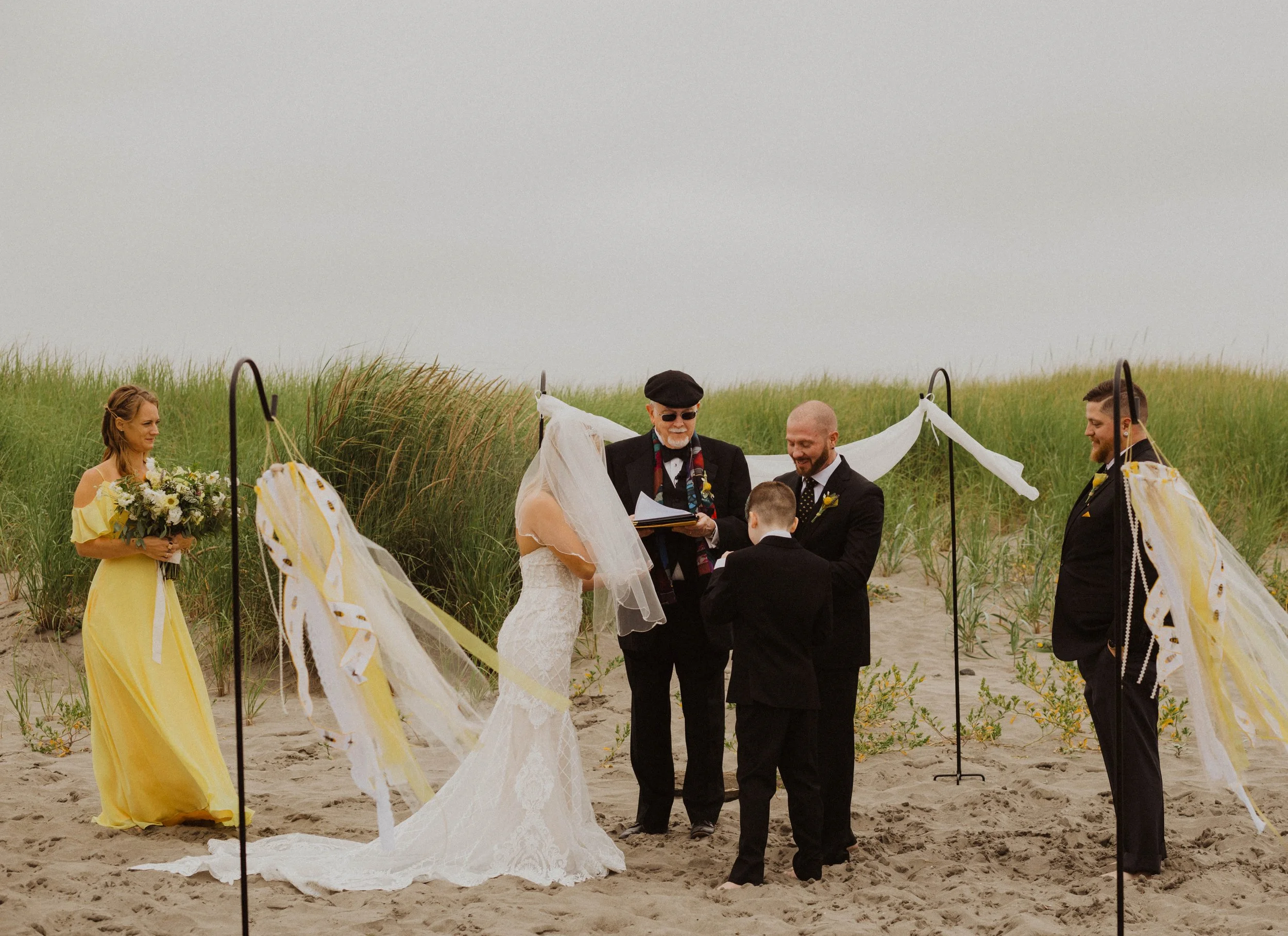 A beach wedding ceremony with a bride in a white lace wedding dress and veil, a groom in a black suit, a young boy in a black suit, and an officiant holding a book, surrounded by wedding party members on a sandy beach with grass and overcast sky. Lon