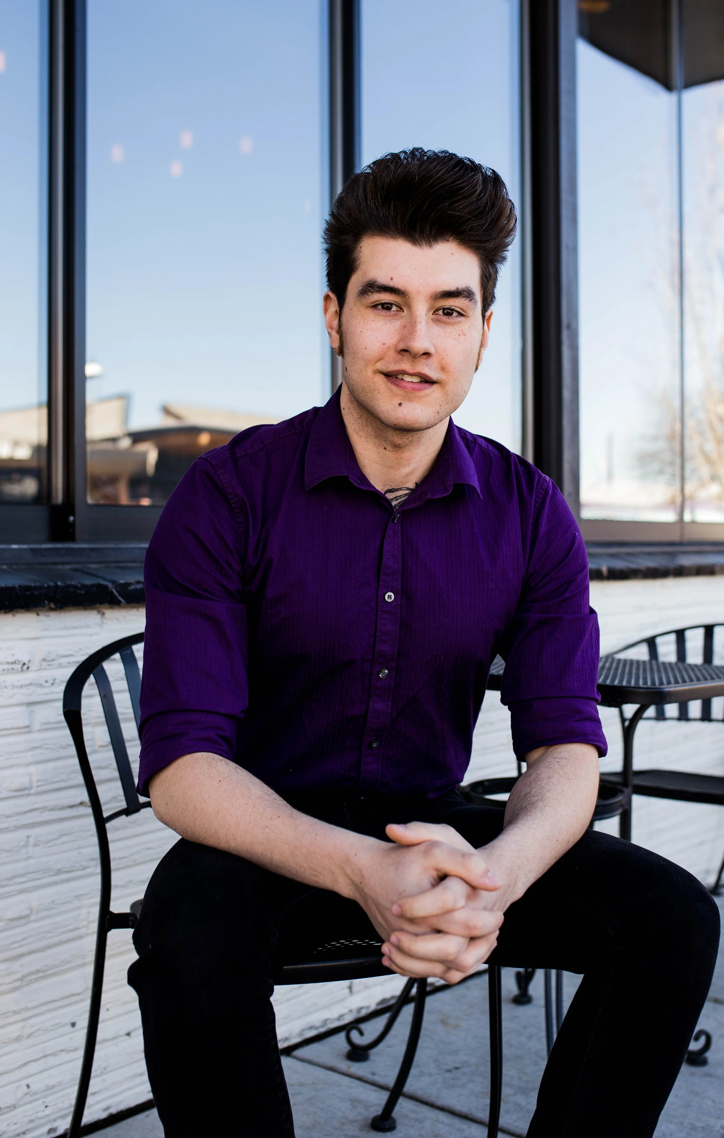 A young man with dark hair, wearing a purple button-up shirt, seated outdoors in front of a large window with reflective glass, sitting on a metal chair with his hands clasped, smiling at the camera. Seattle professional head shot photography