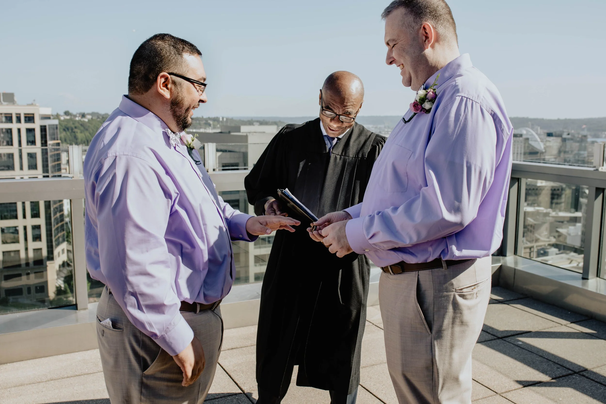 Two men in light purple shirts and khaki pants exchanging vows during a wedding ceremony on a rooftop with a man in a black robe officiating, city skyline in the background. Seattle Municipal Courthouse wedding photography.