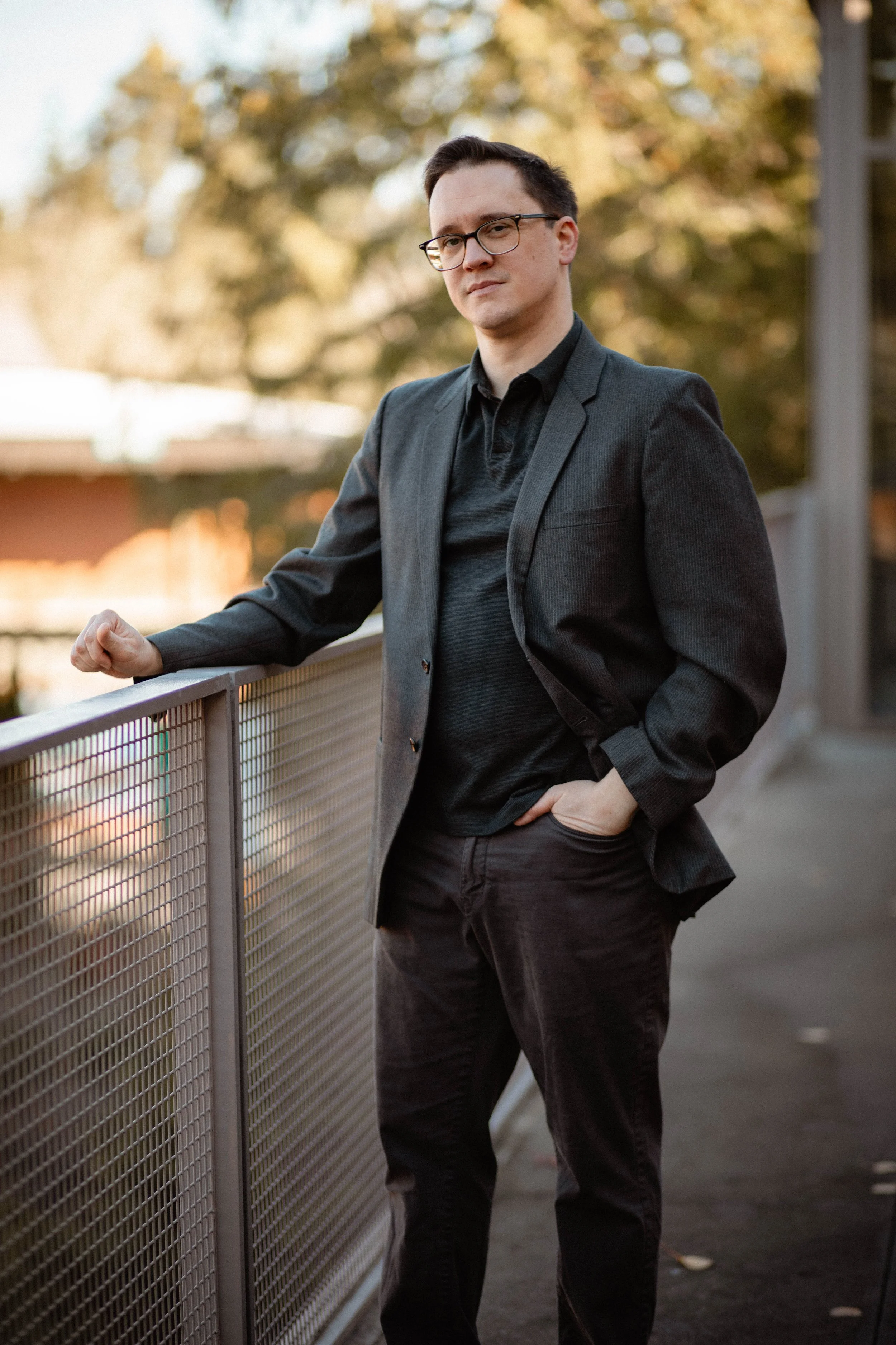 A young man with glasses, wearing a dark blazer and black shirt, standing by a metal railing outdoors during sunset. Seattle professional head shot photography