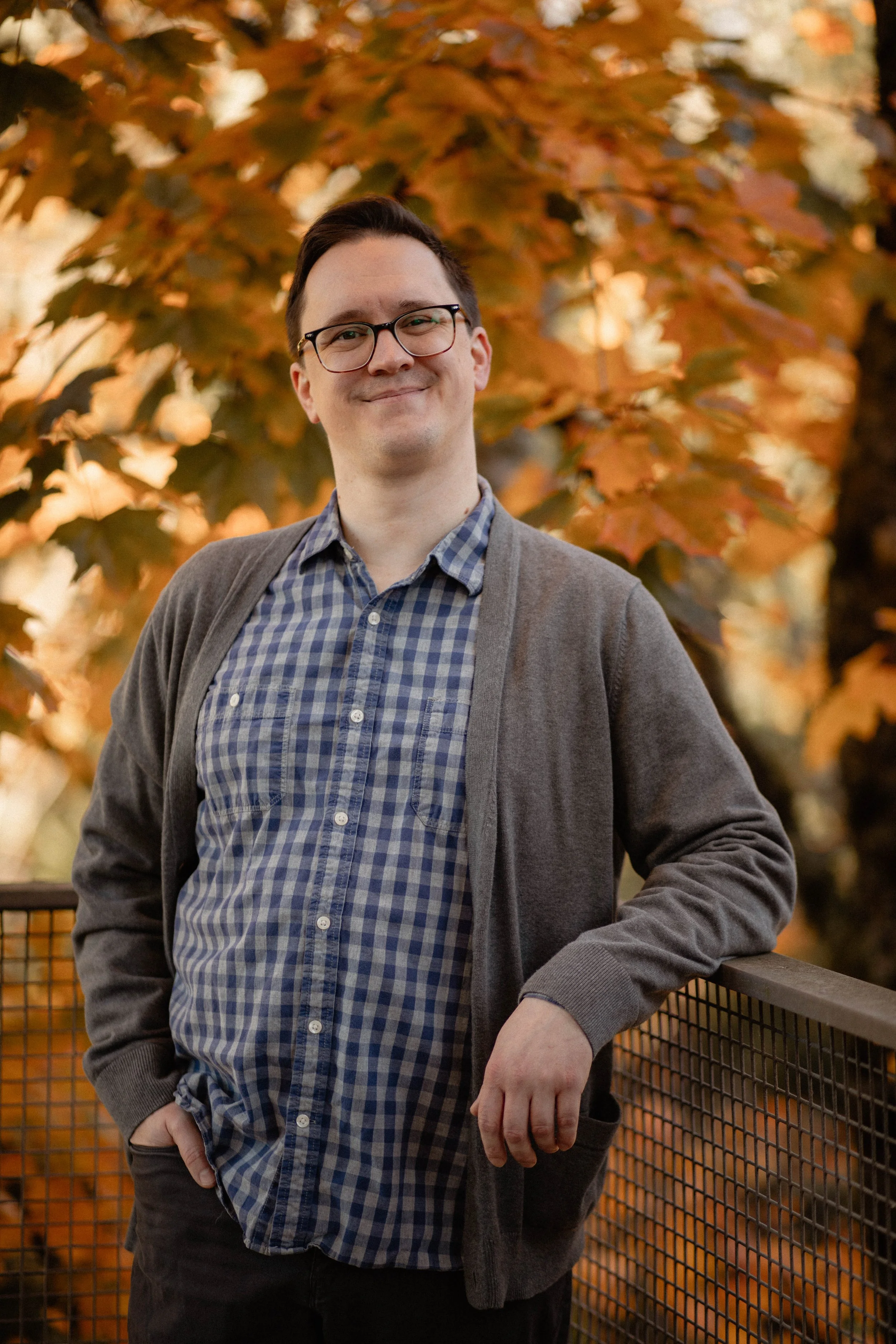 A man in glasses, a blue checked shirt, and a gray cardigan is standing on a bridge with autumn leaves in the background. Seattle professional head shot photography